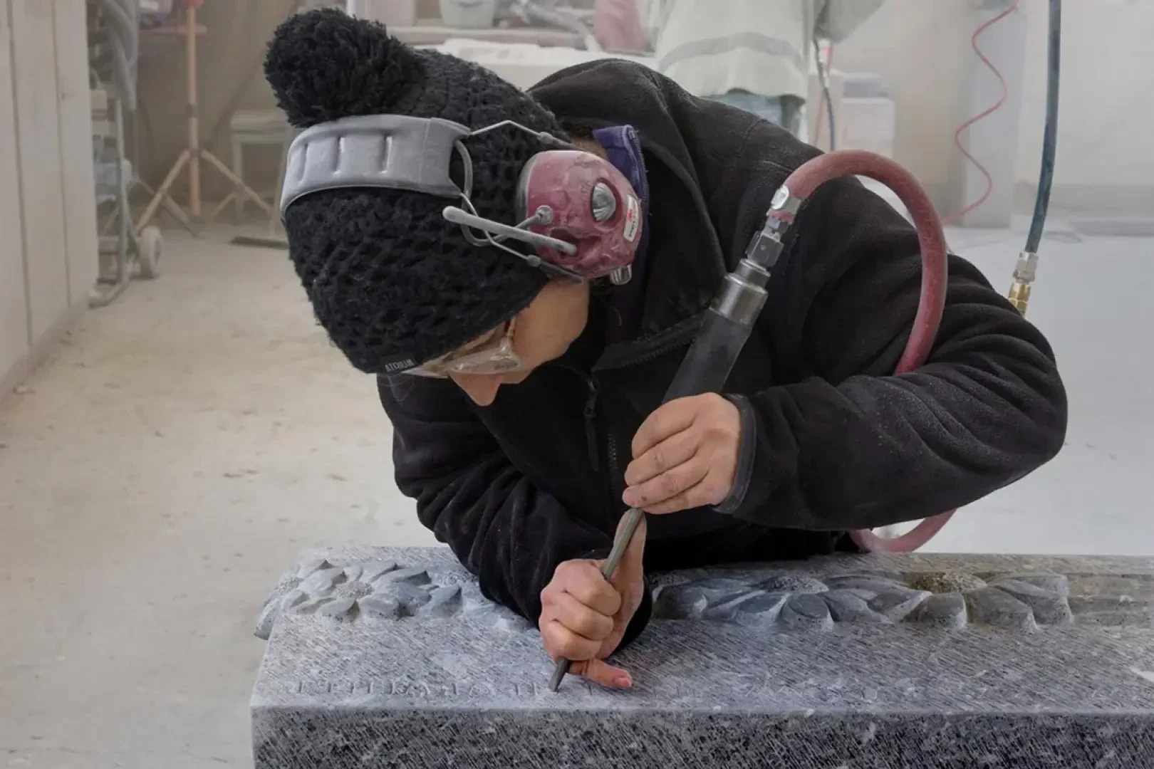Eine Person, die eine schwarze Strickmütze, einen Gehörschutz und eine Schutzbrille trägt, schnitzt mit einem Elektrowerkzeug ein dekoratives Muster in eine Steinplatte in einer Werkstatt.