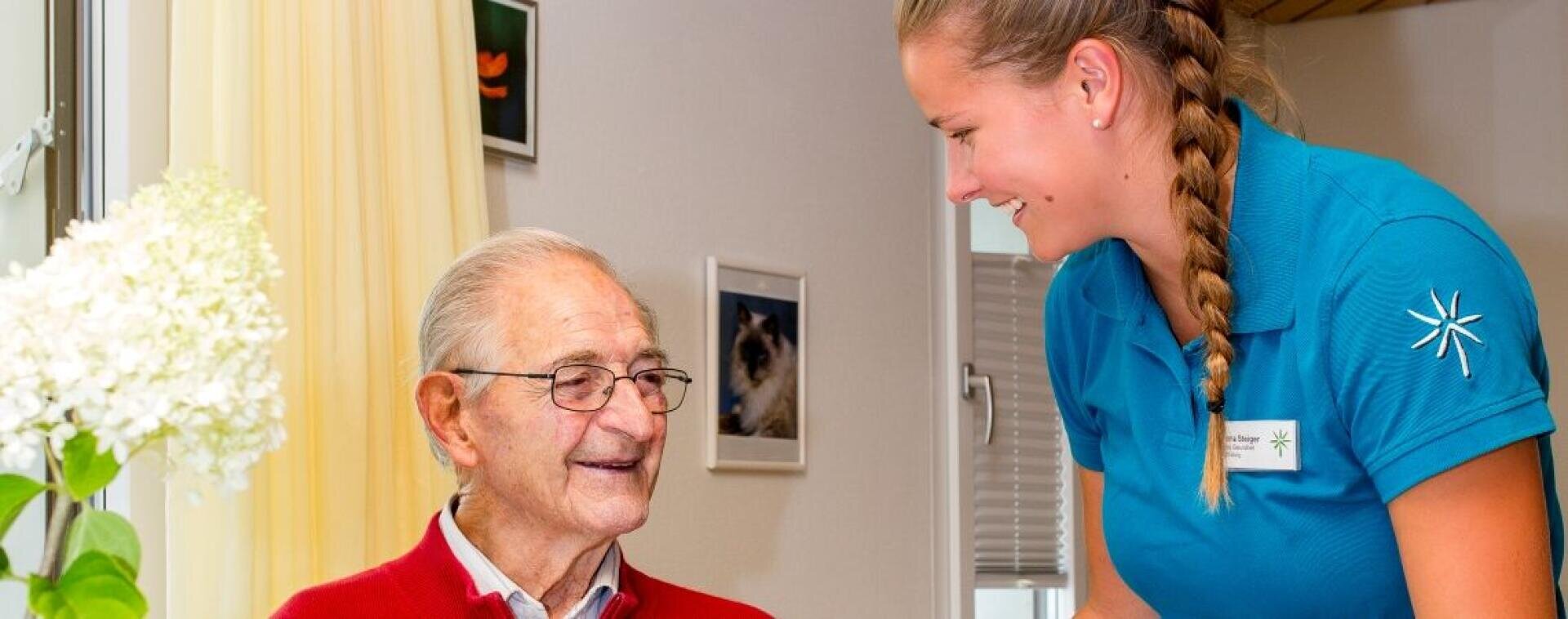 Ein lächelnder älterer Mann in einem roten Pullover sitzt drinnen, während eine junge Frau in einem blauen Hemd mit einem Namensschild lächelnd neben ihm steht. Im Vordergrund ist eine weiße Blume zu sehen, und hinter den beiden ist ein Wandbild zu erkennen.