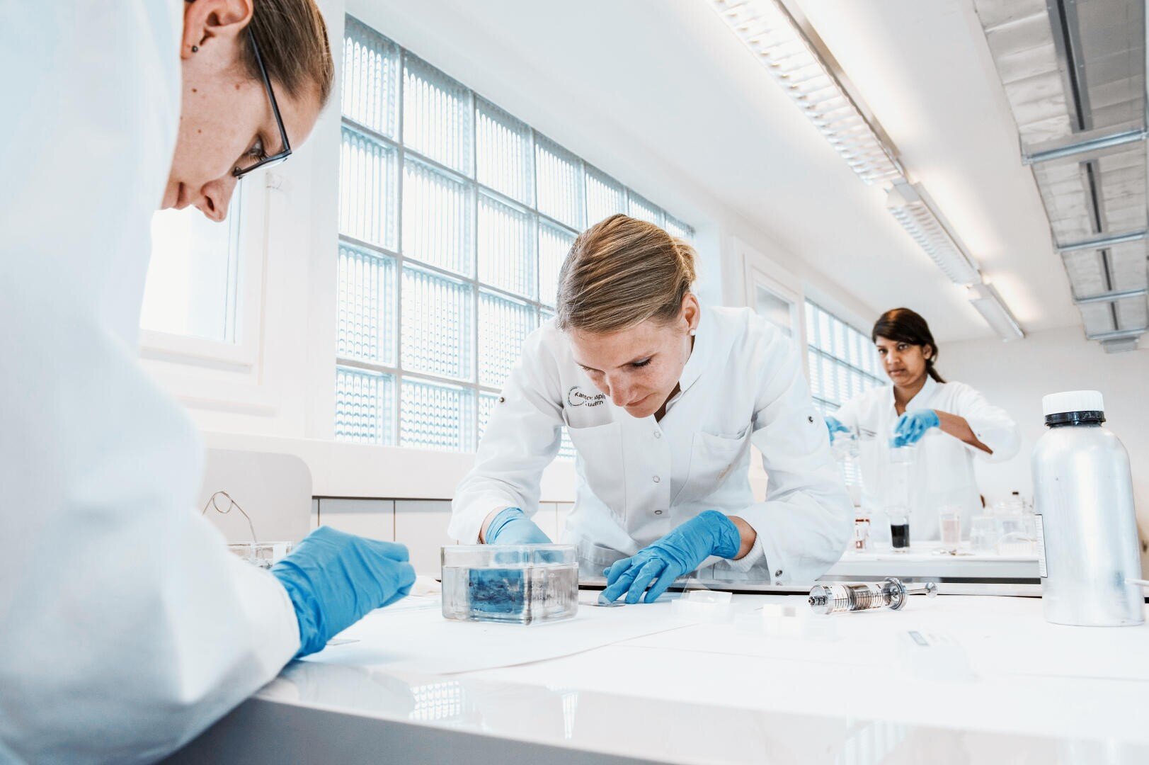 Drei Wissenschaftler in weißen Laborkitteln und blauen Handschuhen arbeiten an einem Labortisch mit verschiedenen Geräten. Die Umgebung ist hell mit großen Fenstern und fluoreszierender Beleuchtung.