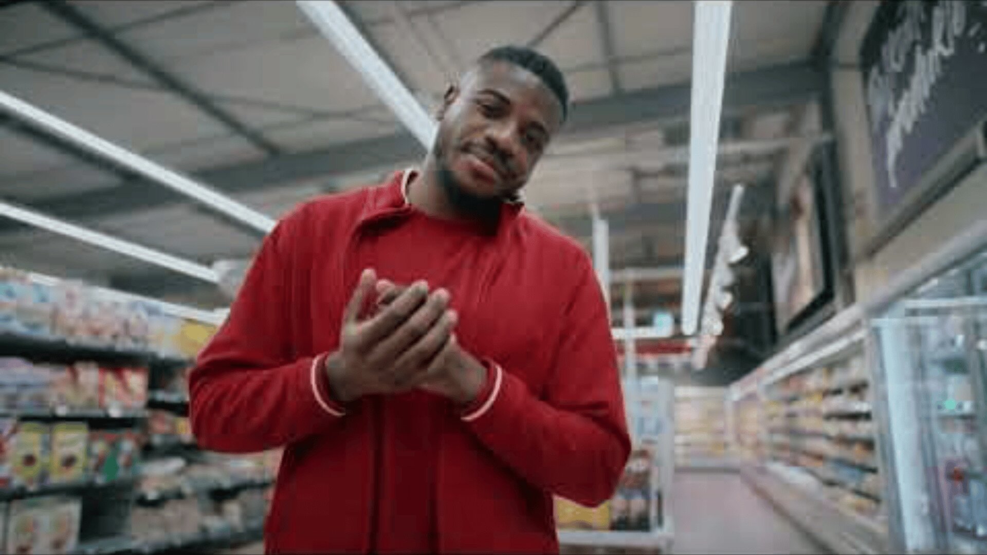 Ein Mann in einer roten Jacke steht in einem Supermarktgang, faltet seine Hände und lächelt leicht. Im Hintergrund sind mit Produkten bestückte Regale zu sehen.