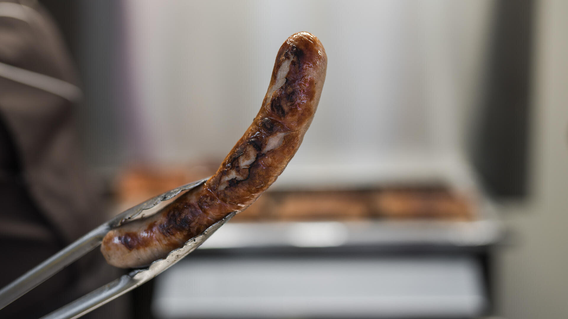 Eine Nahaufnahme einer Metallzange, die eine gegrillte Wurst hält, mit einem verschwommenen Hintergrund, der einen Grill oder eine Kochstelle zeigt.