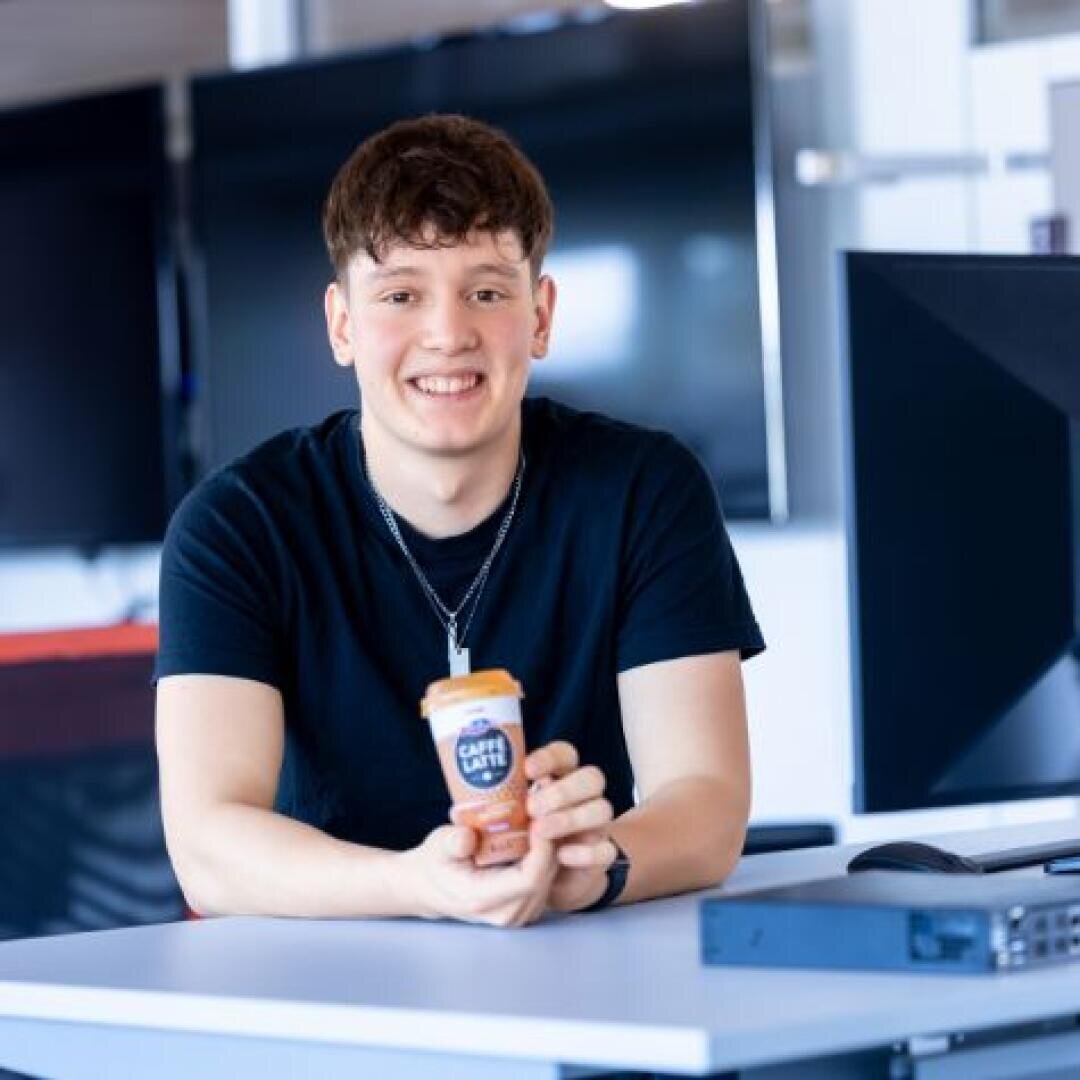 Ein junger Mann mit kurzen braunen Haaren, der ein schwarzes T-Shirt und eine Halskette trägt, sitzt an einem Schreibtisch in einem Büro, hält ein Café Latte Getränk in der Hand und lächelt in die Kamera. Im Hintergrund sind Computermonitore zu sehen.