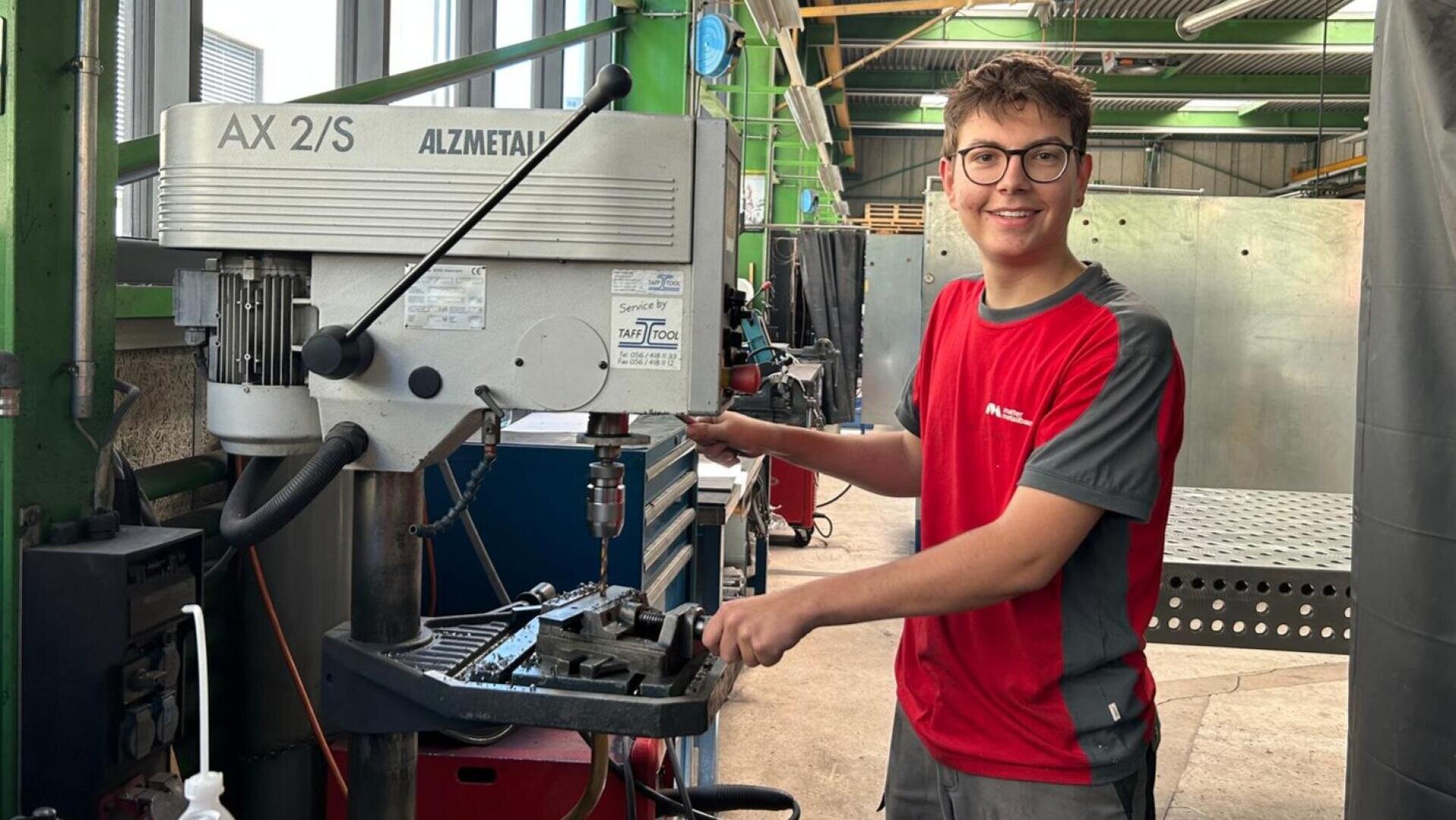 Ein junger Mann mit Brille und einem rot-grauen Hemd bedient in einer Werkstatt eine Metallbohrmaschine.