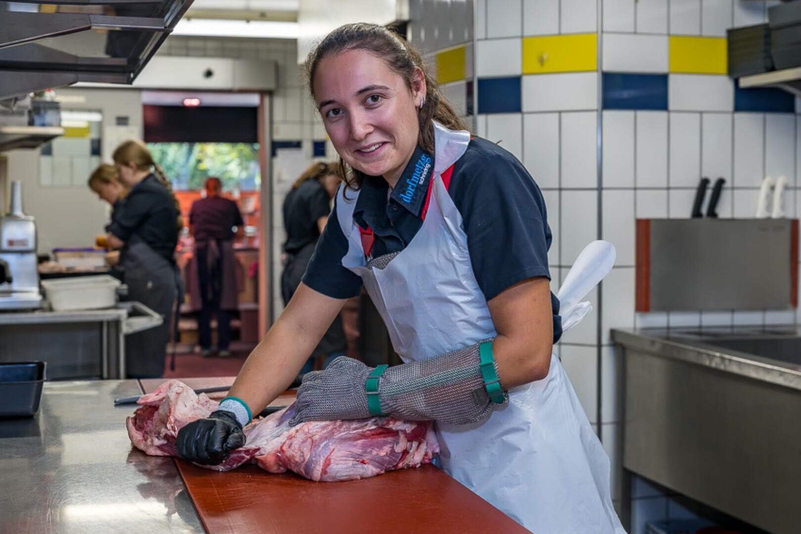 Eine lächelnde Frau, die eine weiße Schürze und einen Sicherheitshandschuh aus Metallgewebe trägt, schneidet auf einer Küchenarbeitsplatte ein großes Stück rohes Fleisch.