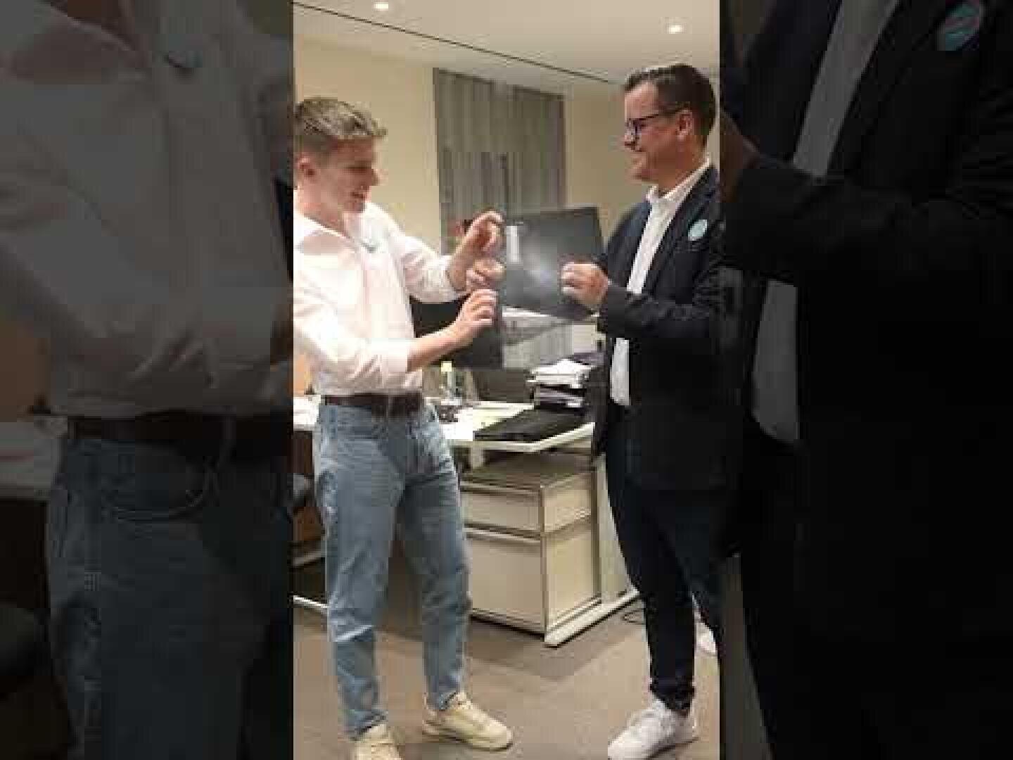Zwei Männer stehen in einem Büro, lächeln und halten einen kleinen Gegenstand in der Hand. Einer trägt ein weißes Hemd und Jeans, der andere eine dunkle Jacke und weiße Turnschuhe. Im Hintergrund sind ein Schreibtisch und ein Bürostuhl zu sehen.