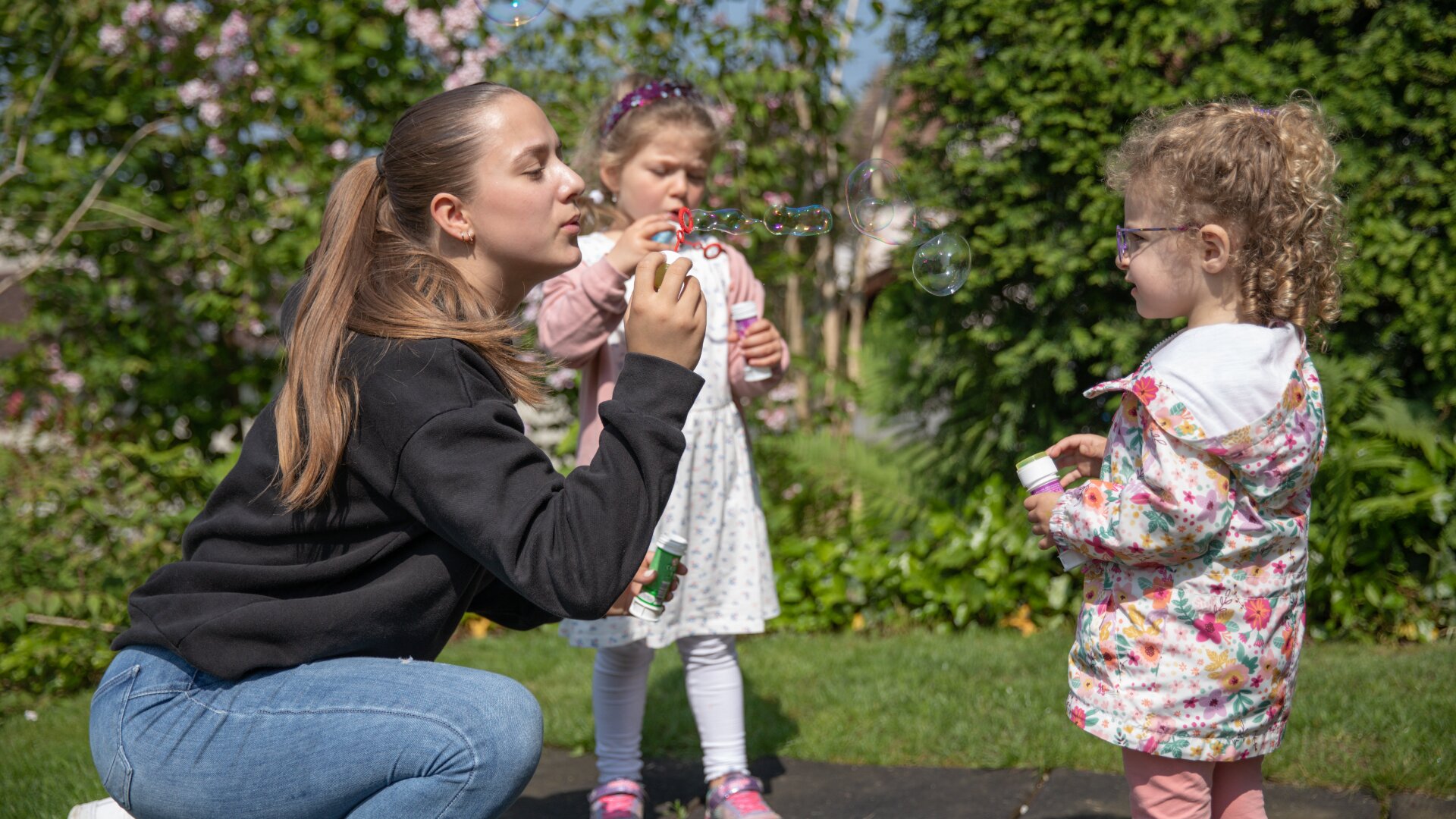 Eine junge Frau kauert und bläst Seifenblasen, während zwei junge Mädchen, eines in einem Kleid und das andere in einer geblümten Jacke, an einem sonnigen Tag im Grünen spielen und zusehen.