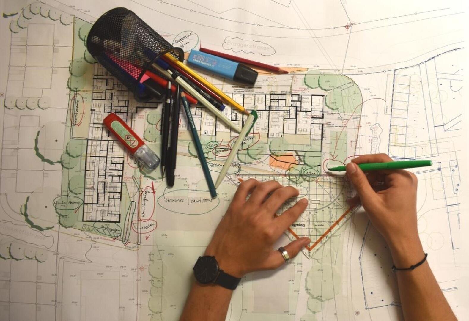 Eine Person zeichnet mit Buntstiften und Markern einen architektonischen Lageplan. Verschiedene Zeichenwerkzeuge wie Stifte, Textmarker und ein Federmäppchen sind auf dem Papier verstreut.