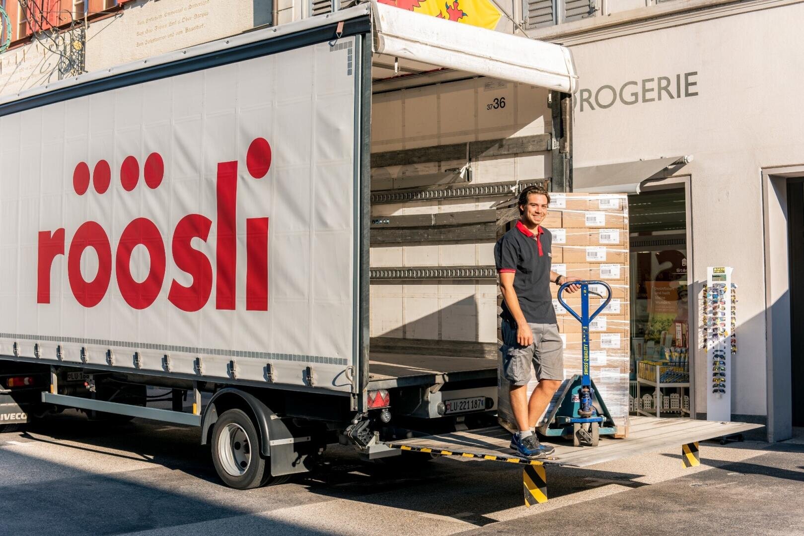 An einem sonnigen Tag steht ein Lieferant auf der Ladefläche eines mit „röösli“ beschrifteten Lastwagens, neben einer Palette mit Kartons und einem blauen Hubwagen, auf einer Rampe vor einem Geschäft.