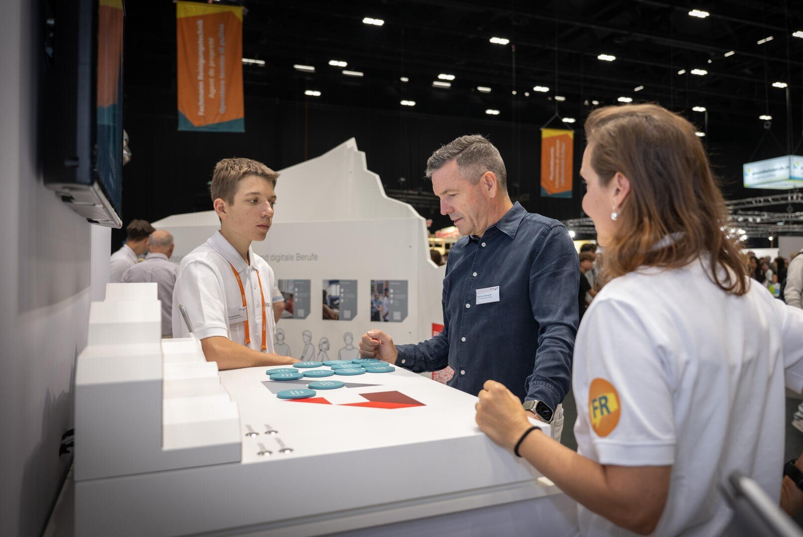 Drei Personen stehen an einem Stand bei einer belebten Veranstaltung in einer Halle und führen ein Gespräch. Zwei tragen weiße Hemden mit orangefarbenen Details, die dritte Person ein blaues Hemd. Auf dem Tisch vor ihnen liegen Broschüren und ein weißes Display.