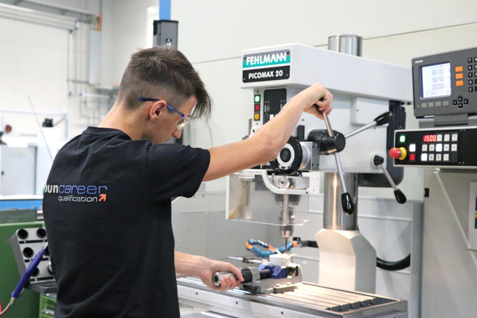 Ein junger Mann mit Schutzbrille arbeitet in einer Werkstatt an einer FEHLMANN PICOMAX 20 Fräsmaschine. Mit einer Hand stellt er die Bedienelemente ein, mit der anderen hält er ein Werkzeug.