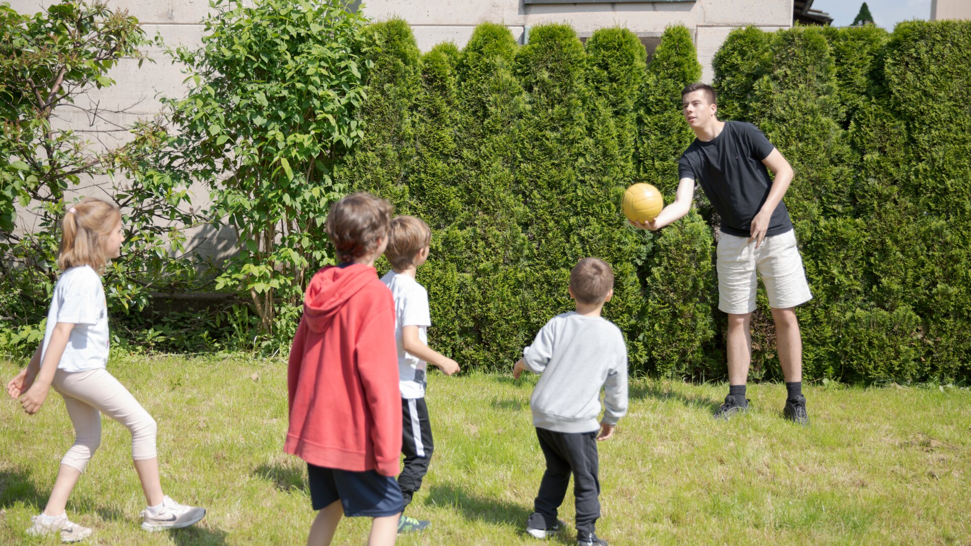 Ein Mann in schwarzem Hemd und kurzen Hosen steht auf dem Rasen und reicht einer Gruppe von vier Kindern einen gelben Ball. Die Kinder schauen ihm aufmerksam zu. Sie befinden sich im Freien, in der Nähe einer grünen Hecke und einer beigen Mauer.