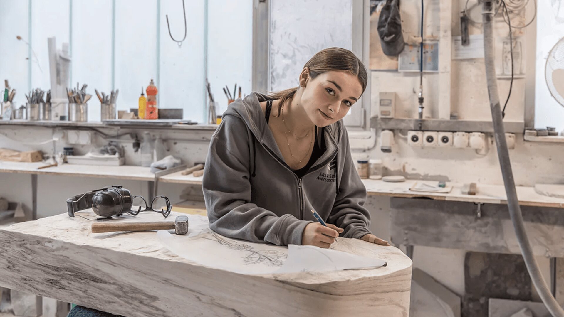 Eine Frau in einer Werkstatt beugt sich über ein großes Stück Holz und skizziert einen Entwurf auf Papier. Sie trägt einen grauen Kapuzenpulli und ist umgeben von Werkzeugen und Kunstzubehör auf Regalen und Tresen.