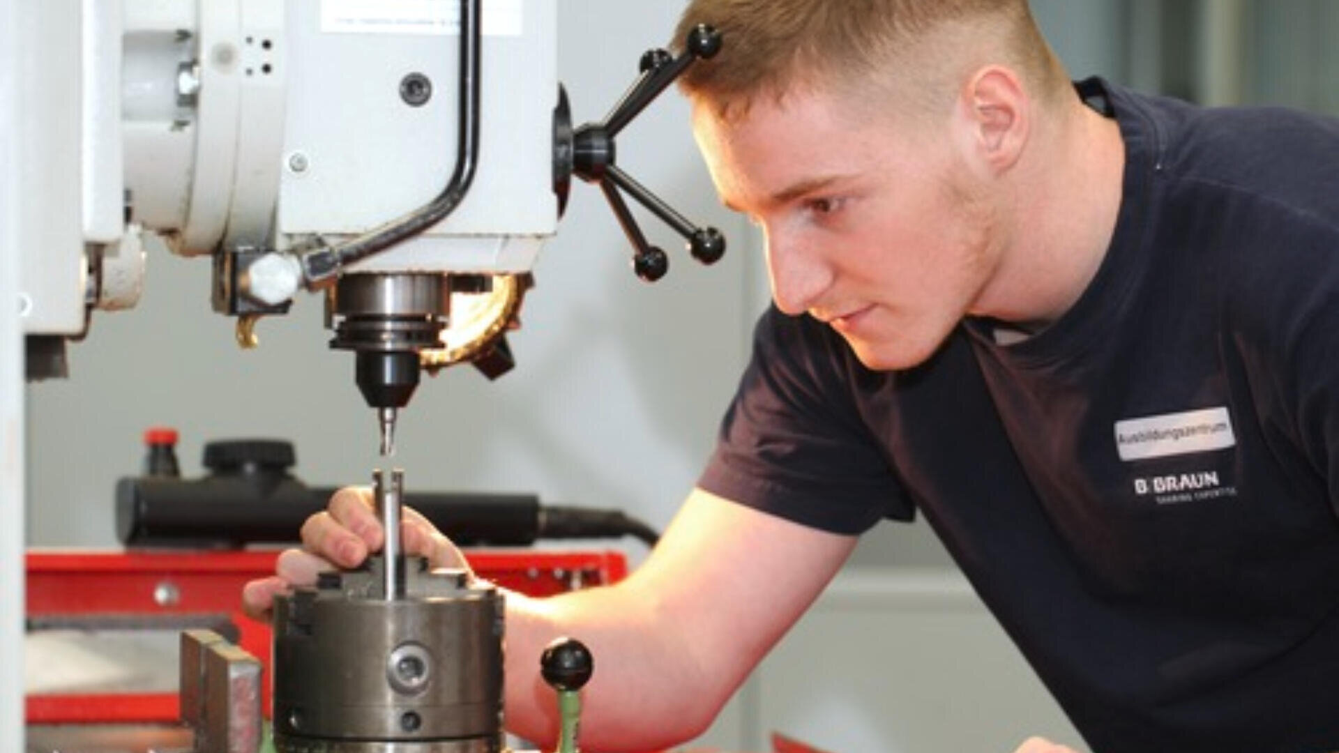 Ein junger Mann bedient eine Präzisionsfräsmaschine und justiert sorgfältig ein Metallteil unter dem Bohrer. Er trägt ein marineblaues Hemd und konzentriert sich auf seine Aufgabe in einer gut beleuchteten Werkstatt.