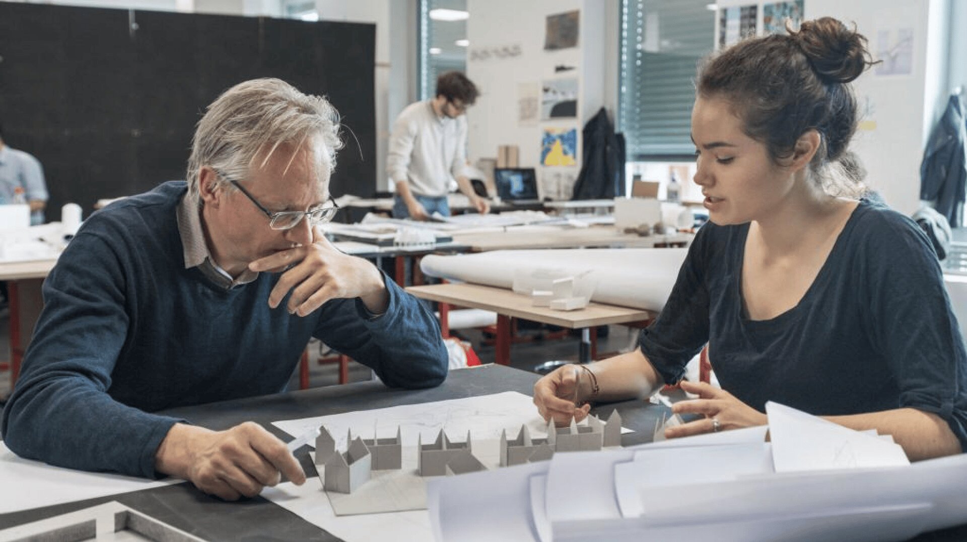 Ein älterer Mann und eine junge Frau diskutieren an einem Tisch in einem Designstudio über ein Architekturmodell, um sie herum liegen Papiere und Modelle. Im Hintergrund arbeiten andere Personen.