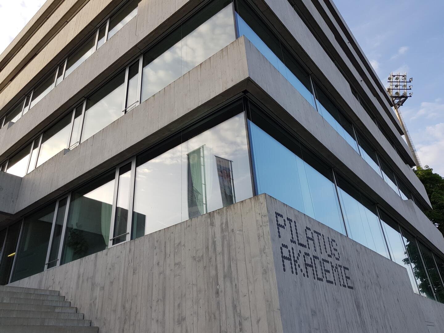 Modernes Betongebäude mit großen Glasfenstern, in denen sich der Himmel und die Wolken spiegeln. Der Schriftzug PILATUS AKADEMIE ist an der Außenwand angebracht. Eine Treppe führt hinauf zum Eingang auf der linken Seite.