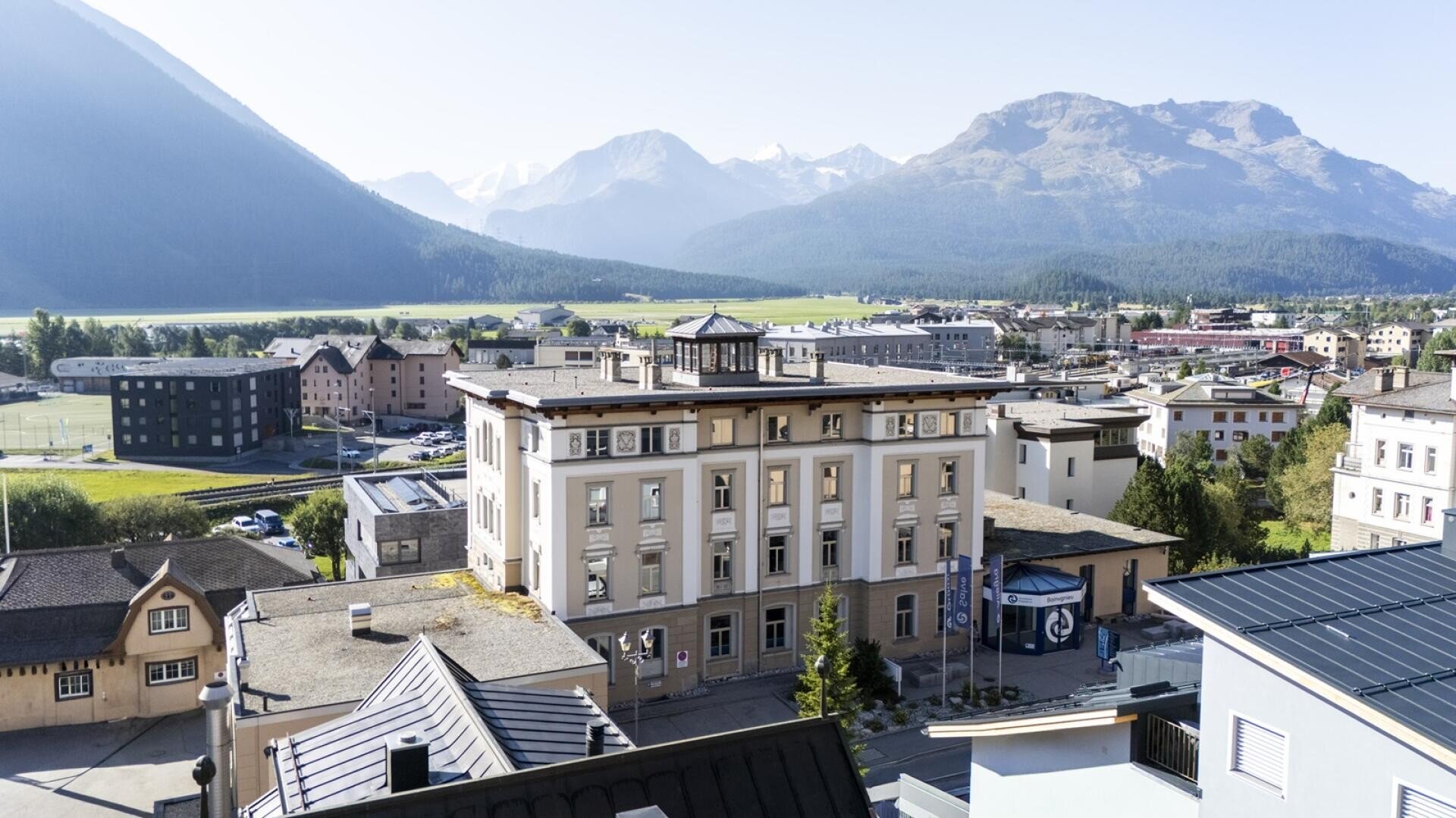 Luftaufnahme von Campus in Samedan mit historischen Gebäuden, gelegen in einem Tal, umgeben von grünen Feldern und sicht auf den Piz Palü und Piz Bernina unter einem strahlend blauen Himmel.