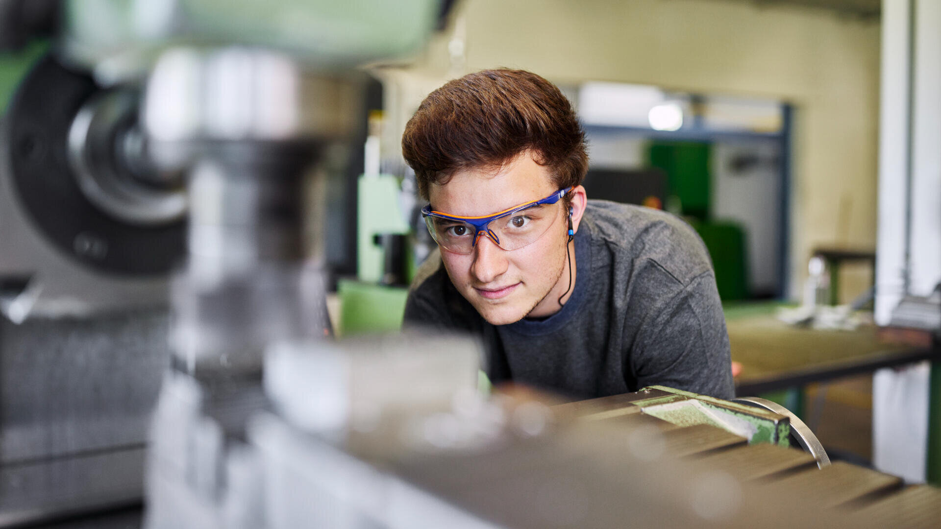 Ein junger Mensch mit einer Schutzbrille beobachtet eine Maschine in einer Werkstatt oder in der Produktion und konzentriert sich auf die laufenden Geräte.