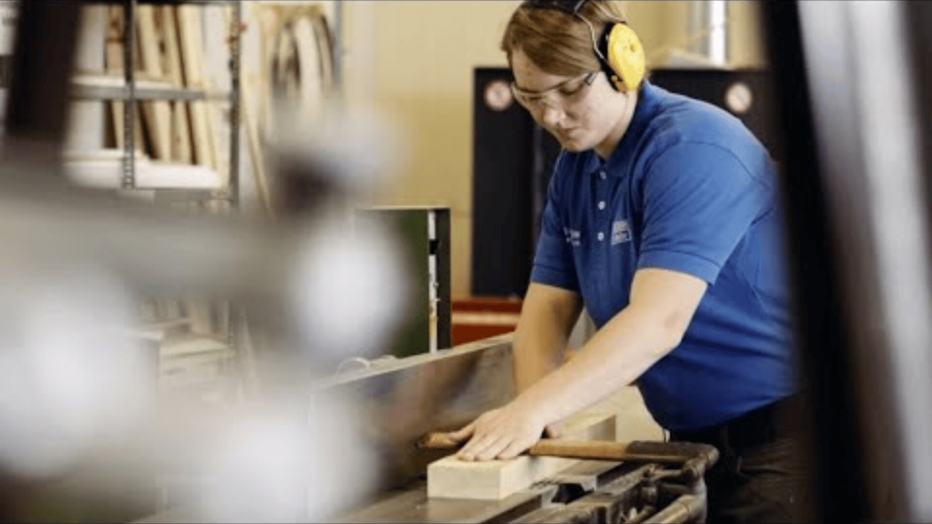 Eine Person, die eine Schutzbrille und einen Gehörschutz trägt, bedient eine Holzbearbeitungsmaschine und führt ein Holzstück durch die Maschine in einer Werkstatt. Im Hintergrund sind Regale mit Materialien zu sehen.