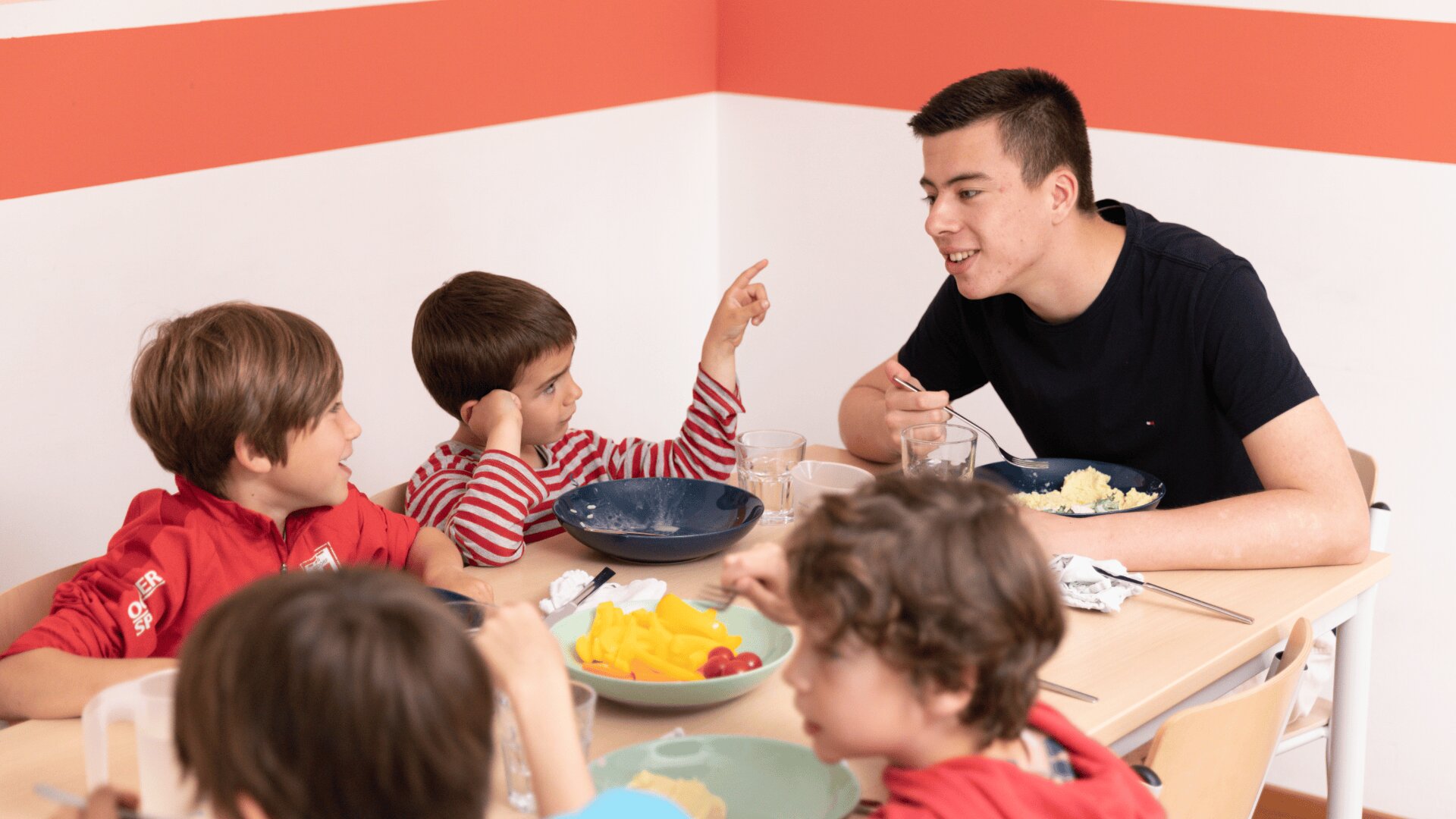 Eine Gruppe kleiner Kinder und ein Erwachsener sitzen um einen Tisch, essen und unterhalten sich. Die Kinder scheinen beschäftigt zu sein, wobei ein Kind auf etwas zeigt, während der Erwachsene lächelt. Auf dem Tisch stehen Teller mit Essen und Getränken. Die Umgebung ist hell und fröhlich.