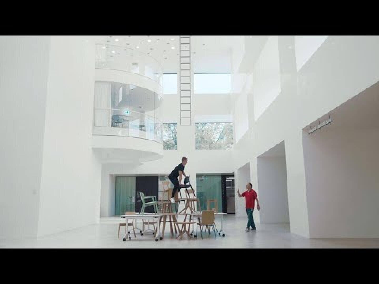 Eine Person steht auf einem hohen, instabilen Stapel von Stühlen und Tischen in einem großen, modernen weißen Raum, während eine andere Person in einem roten Hemd aus der Nähe zusieht.