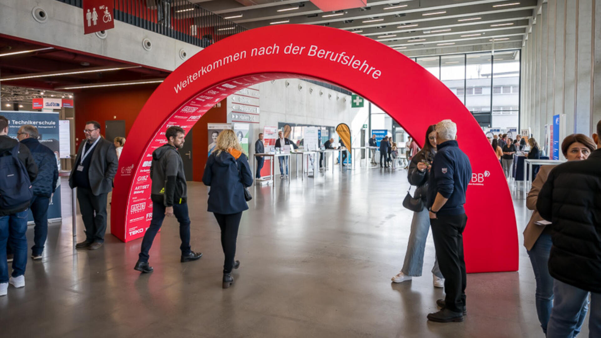 Menschen mischen sich in einem modernen Innenraum bei einer Veranstaltung und gehen unter einem großen roten Bogen mit weißer deutscher Aufschrift "Weiterkommen nach der Berufslehre" hindurch. Im Hintergrund sind verschiedene Stände zu sehen.