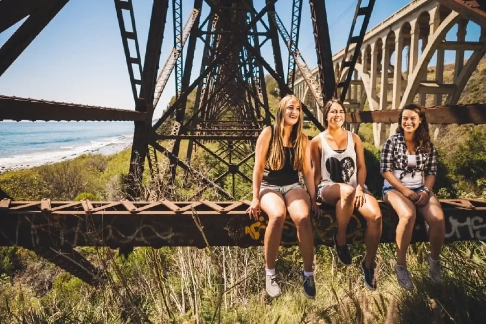 Drei junge Frauen sitzen lächelnd auf einem rostigen Stahlträger einer Eisenbahnbrücke über einer Grünanlage, mit dem Meer und einer weiteren Brücke im Hintergrund an einem sonnigen Tag.