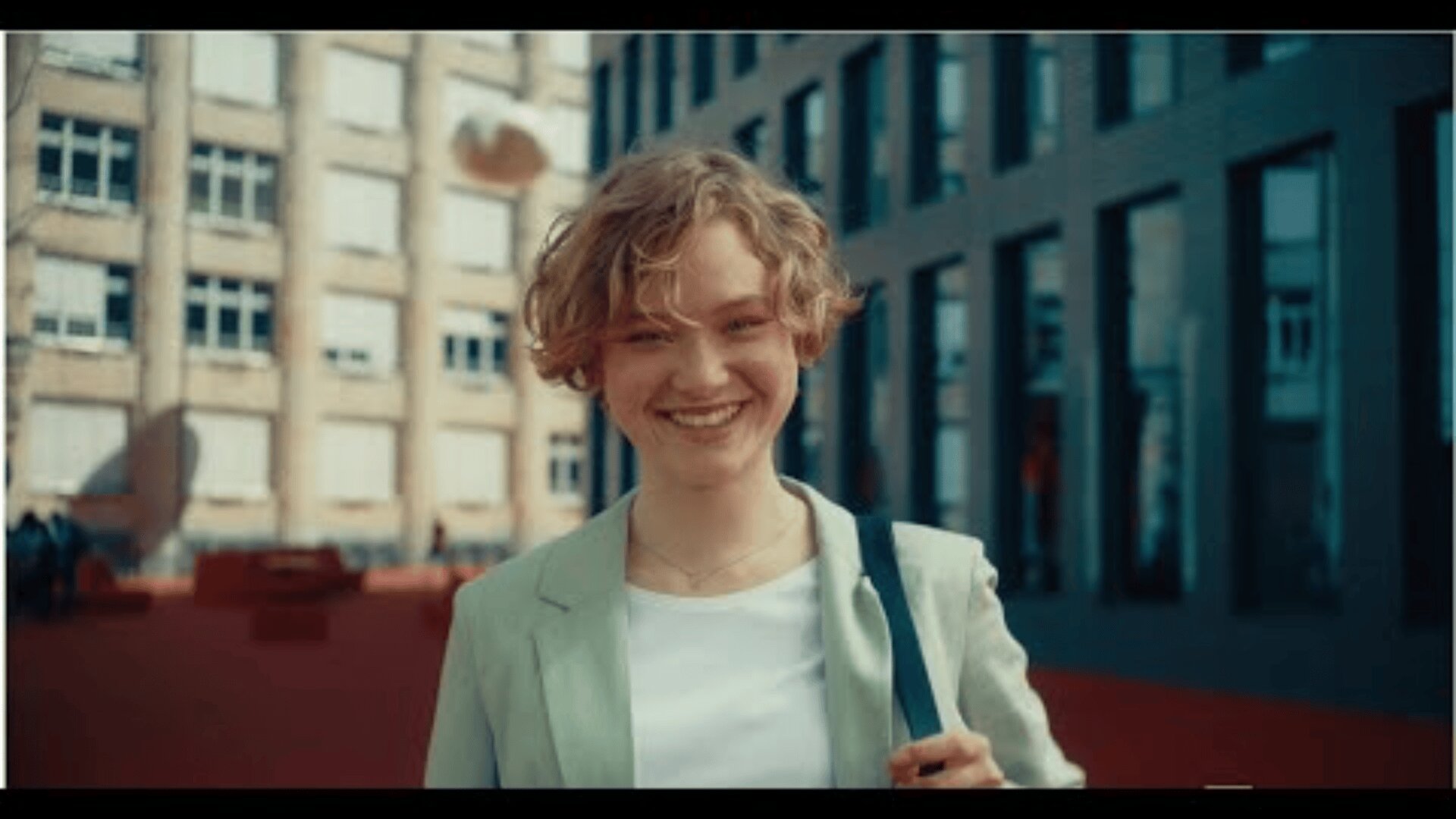 Eine Person mit kurzem, lockigem blondem Haar, die einen hellen Blazer und ein weißes Oberteil trägt, lächelt, während sie im Freien vor einem modernen Gebäude steht.