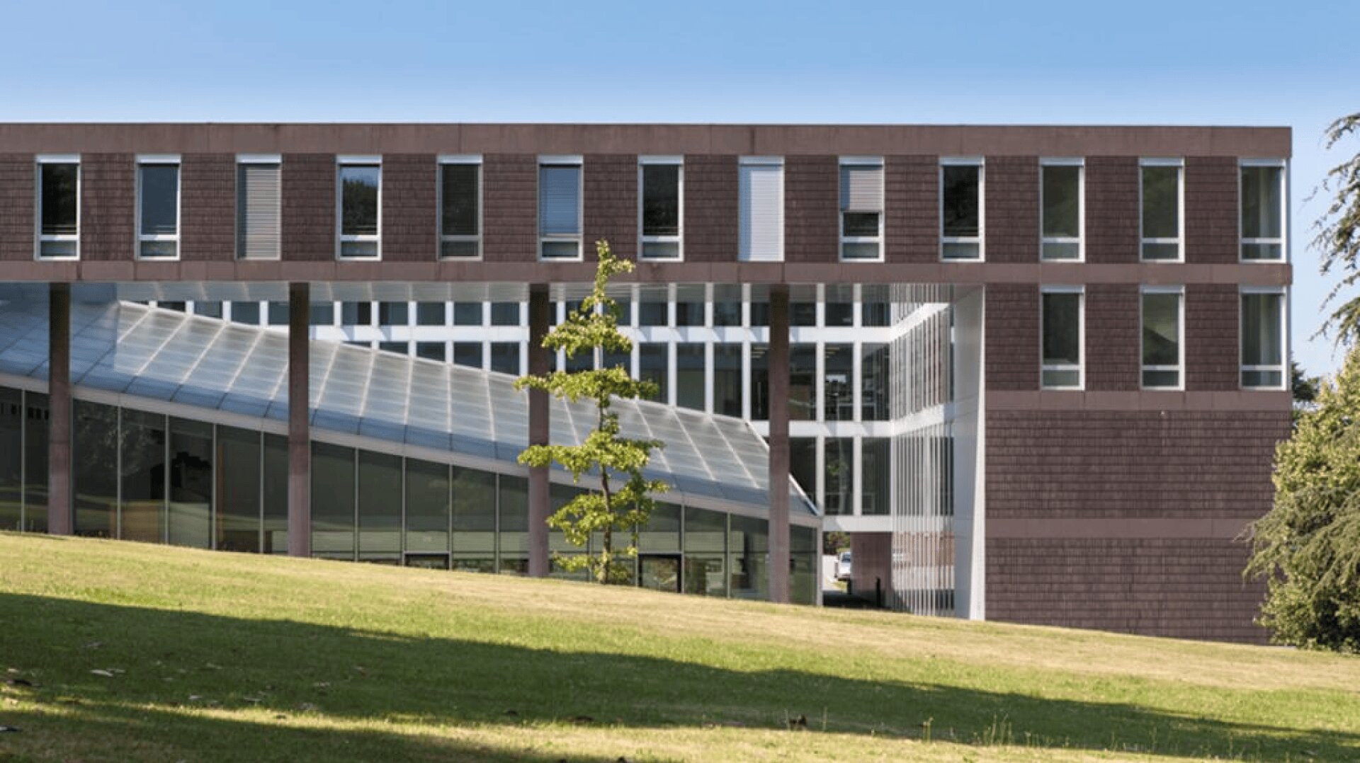 Ein modernes Gebäude mit großen Glasfenstern und einem Flachdach, das mit rechteckigen braunen und grauen Platten versehen ist. Es steht auf einer Rasenfläche mit einigen kleinen Bäumen und einem klaren blauen Himmel im Hintergrund.