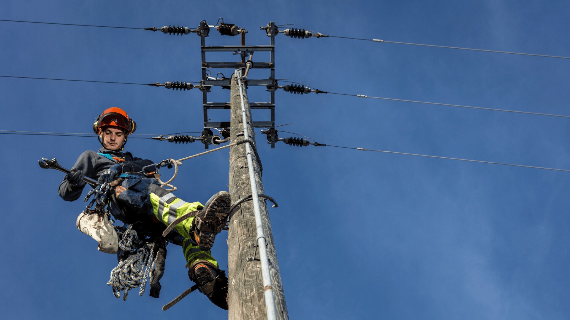 Ein Arbeiter eines Energieversorgungsunternehmens klettert mit Sicherheitsausrüstung auf einen hölzernen Strommast, über dem sich bei strahlend blauem Himmel elektrische Leitungen befinden. Der Arbeiter benutzt zur Sicherheit einen Klettergurt und eine Kletterausrüstung.