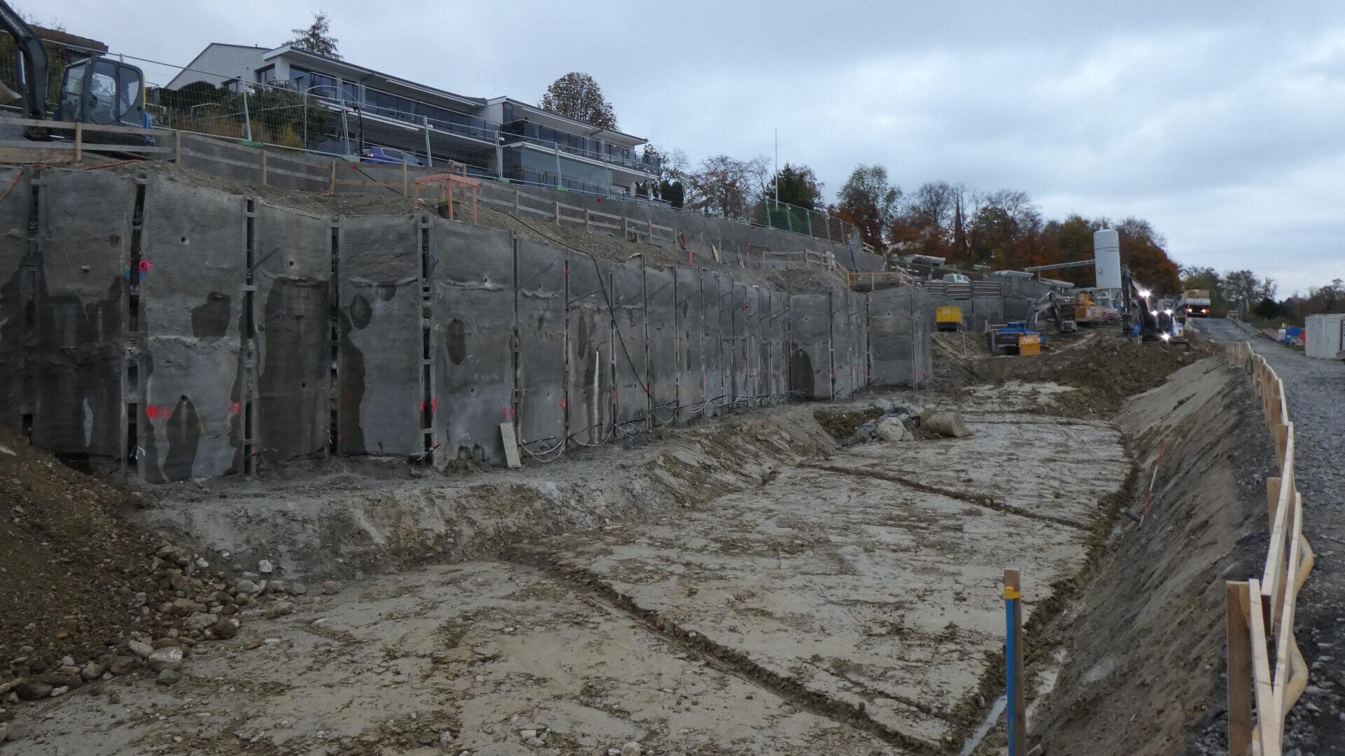 Eine Baustelle mit einer großen, ausgehobenen Fläche und hohen, mit Beton verstärkten Stützmauern. Baumaschinen und -materialien sind zu sehen, im Hintergrund stehen Häuser und Bäume bei bewölktem Himmel.