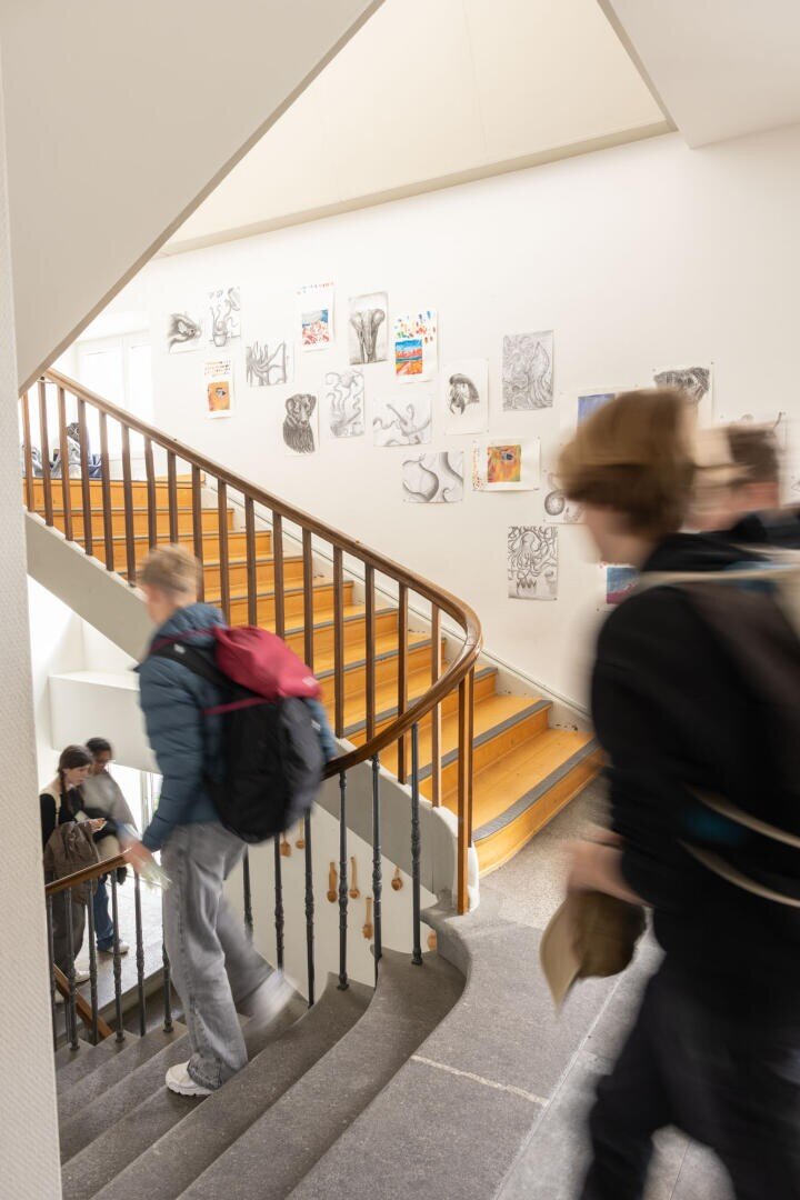 Schüler gehen eine geschwungene Treppe in einem Gebäude auf und ab. An der weißen Wand über der Holztreppe sind Kunstwerke und Zeichnungen ausgestellt. Die Szene wirkt belebt und weist eine gewisse Bewegungsunschärfe auf.