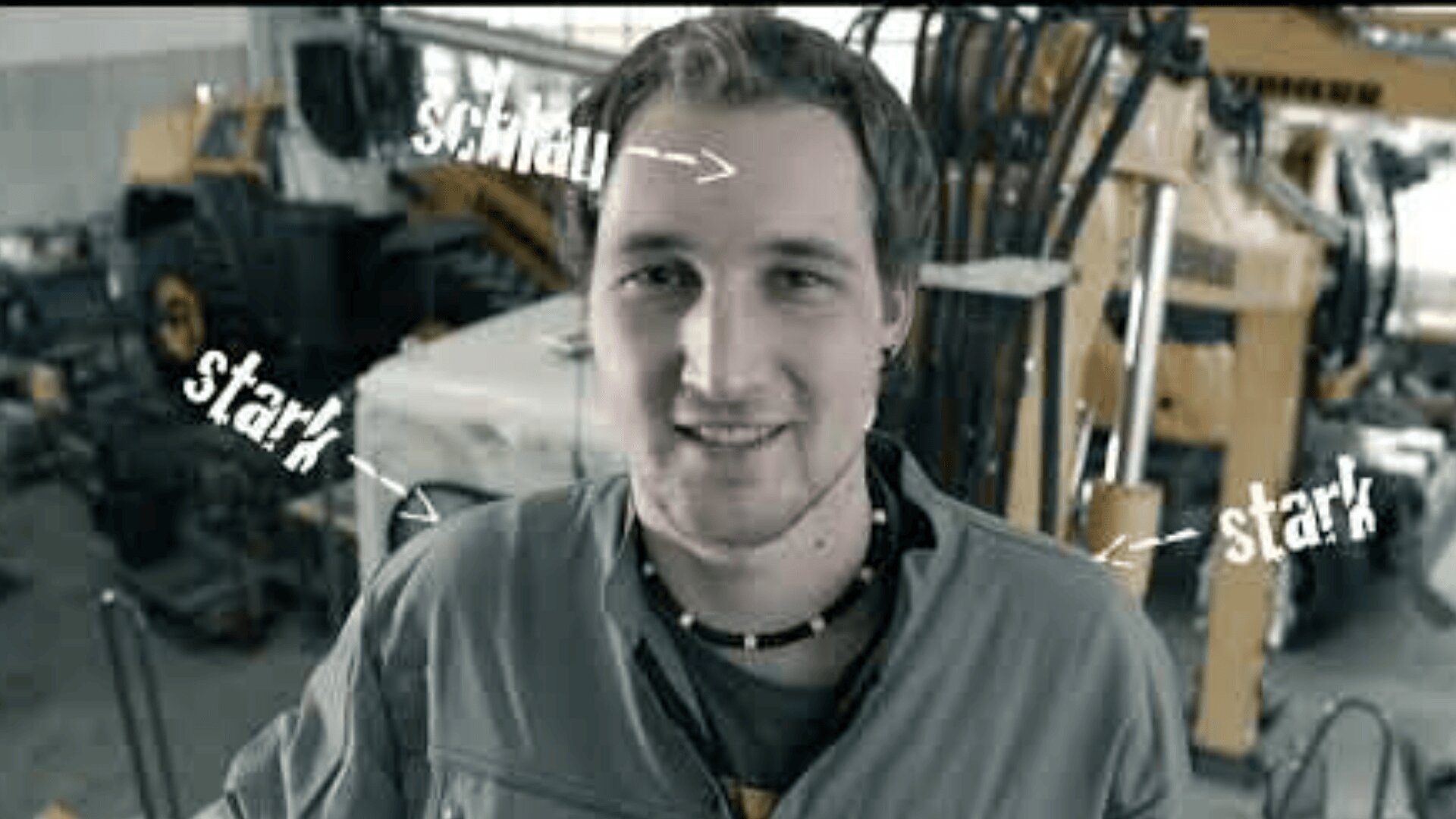 Ein junger Mann in einer Werkstatt steht lächelnd vor der Kamera, umgeben von Baumaschinen. Weiße, handgeschriebene Etiketten mit der Aufschrift "schlau" über seinem Kopf und "stark" auf beiden Seiten.
