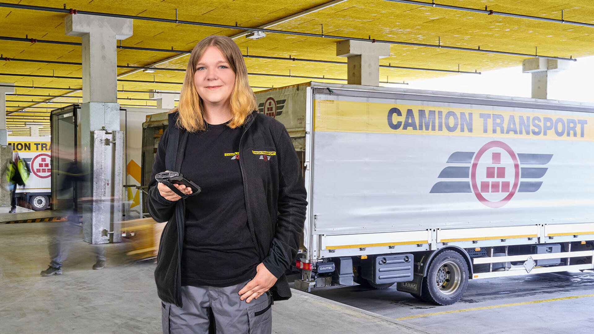 Eine Frau in einer schwarzen Jacke steht vor einem LKW von CAMION TRANSPORT in einer Laderampe. Sie hält ein Handheld-Gerät in der Hand und lächelt, während im Hintergrund andere Arbeiter und Geräte zu sehen sind.