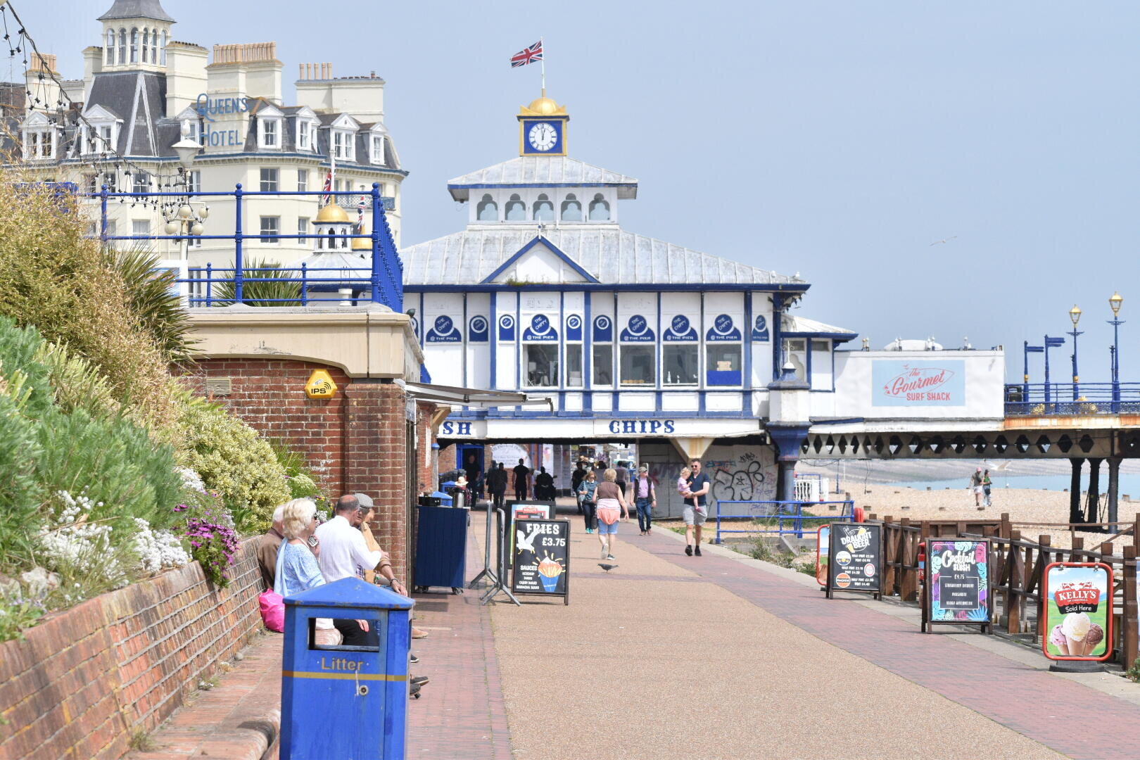 Menschen sitzen auf Bänken entlang einer Strandpromenade neben einem blau-weißen Piergebäude mit Uhrenturm. Auf Schildern wird für Strandgut und Fish and Chips geworben. Im Hintergrund sind der Strand und das Meer zu sehen.