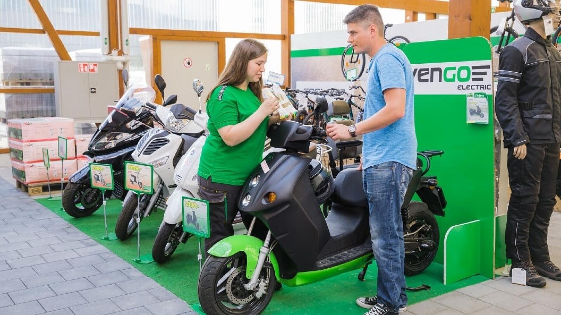Eine Frau in einem grünen Hemd und ein Mann in einem blauen Hemd besprechen einen Elektroroller in einem Ausstellungsraum mit mehreren Rollern und einem grünen "Vengo Electric"-Schild im Hintergrund.