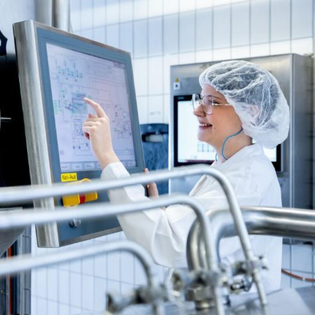 Eine Person im Laborkittel, mit Haarnetz und Brille lächelt und berührt ein großes Touchscreen-Bedienfeld in einer modernen Industrie- oder Laborumgebung mit Metallrohren im Vordergrund.