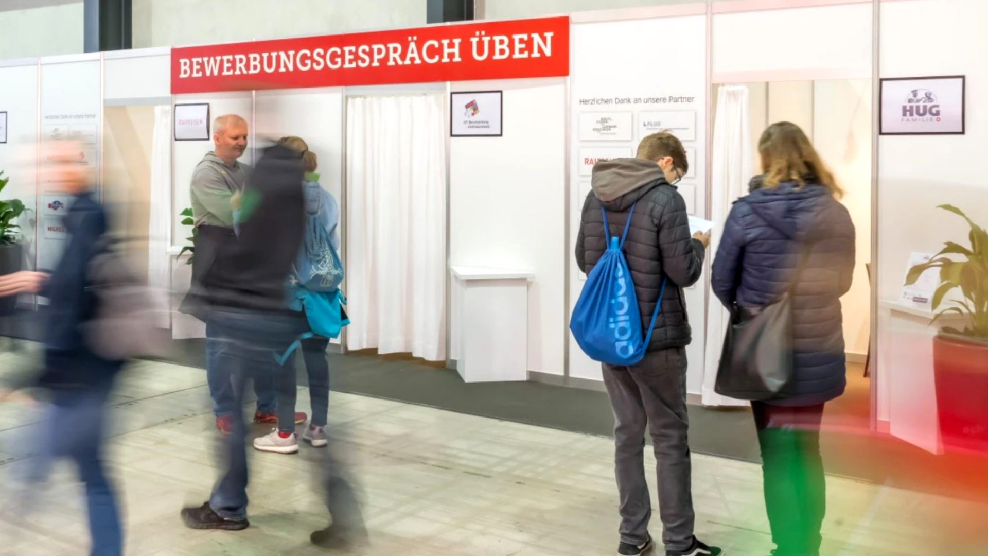 Auf einer Jobmesse stehen Menschen vor Ständen unter einem Schild mit der Aufschrift „Bewerbungsgespräch üben“. Einige unterhalten sich mit den Mitarbeitern an den Ständen, andere warten oder lesen.