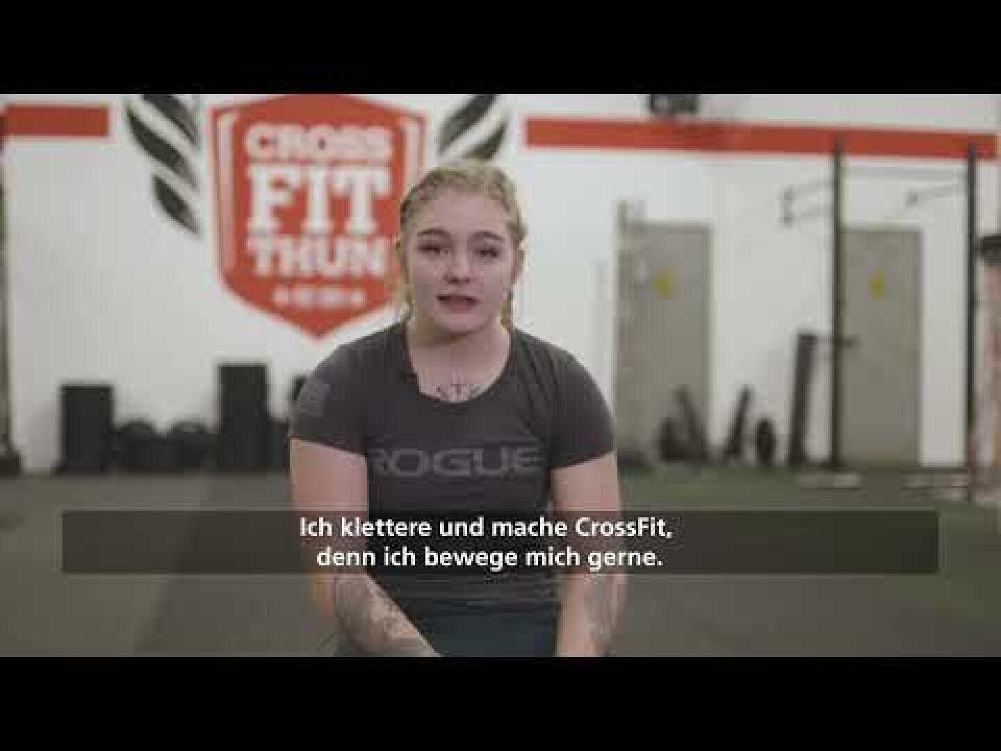 Eine junge Frau sitzt in einem Fitnessstudio mit CrossFit Thun an der Wand hinter ihr. Sie trägt ein dunkles Hemd und hat ihr blondes Haar zurückgebunden. Die deutschen Untertitel lauten: Ich klettere und mache CrossFit, denn ich bewege mich gerne.