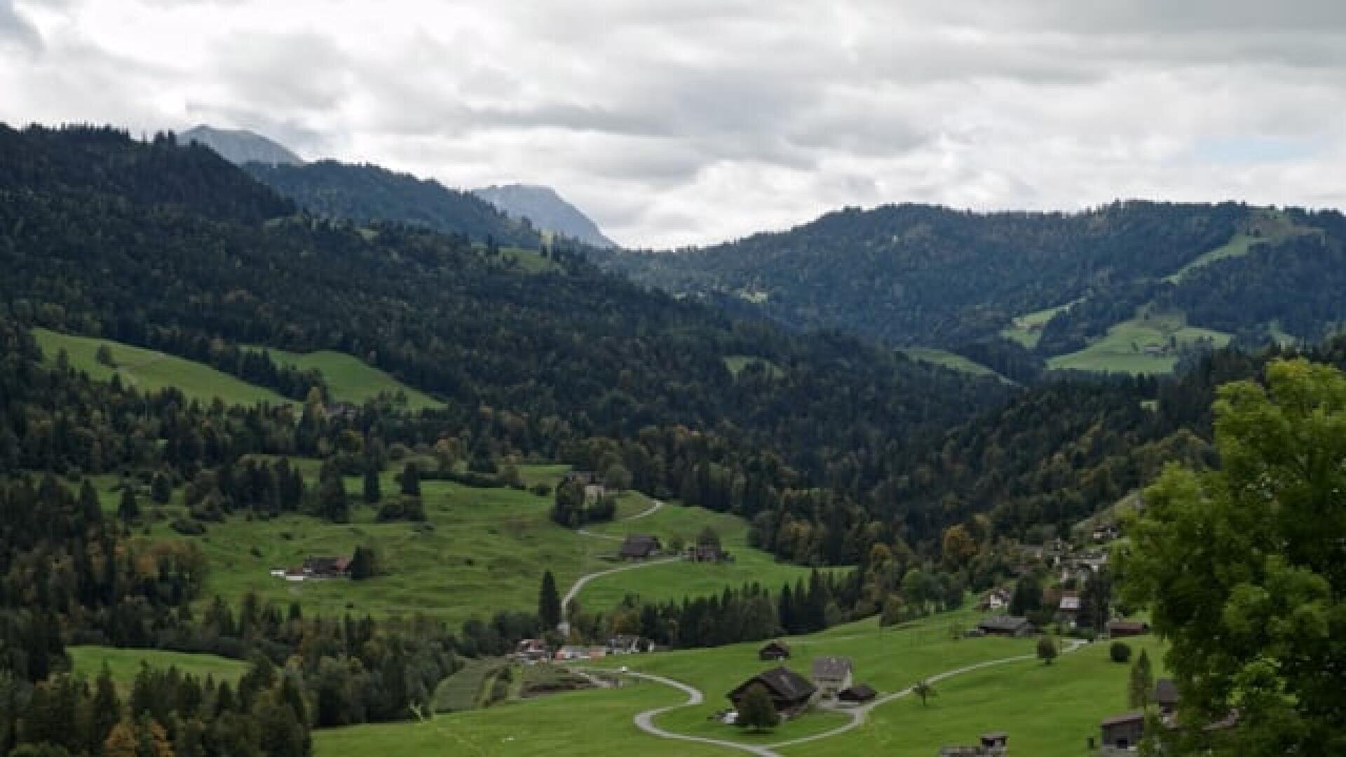 Eine malerische Aussicht auf sanfte grüne Hügel mit Häusern und Bäumen, umgeben von bewaldeten Bergen unter einem bewölkten Himmel.