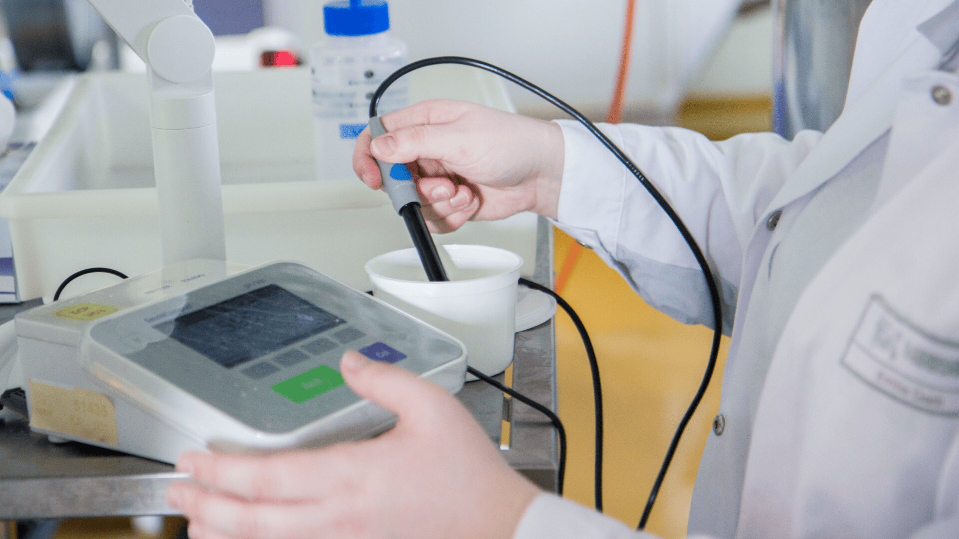 Eine Person in einem Laborkittel verwendet eine tragbare elektronische Sonde, die mit einem digitalen Gerät verbunden ist, und platziert die Sonde in einem weißen Becher auf einem Tisch aus rostfreiem Stahl in einer Laborumgebung.
