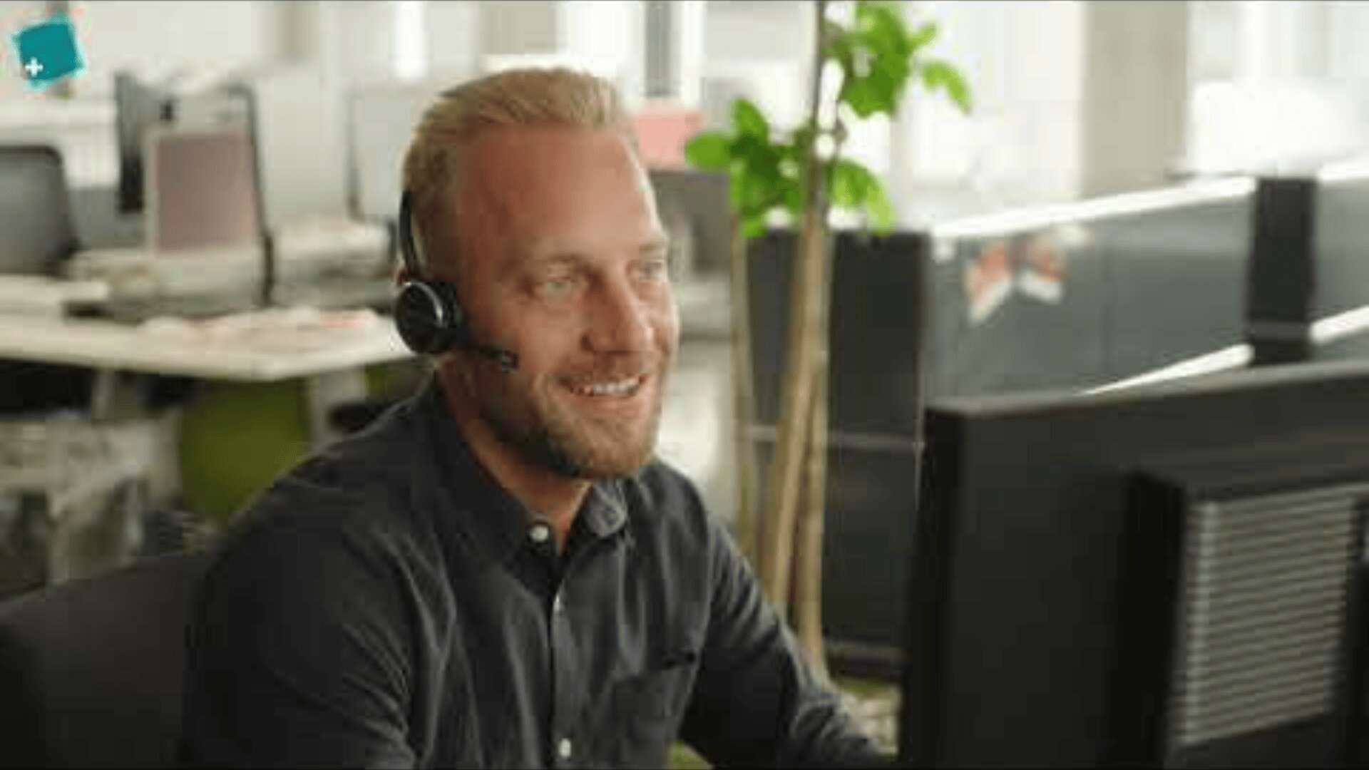 Ein Mann mit einem Headset sitzt an einem Schreibtisch und schaut lächelnd auf einen Computerbildschirm in einem modernen Büro mit Pflanzen und Arbeitsplätzen im Hintergrund.