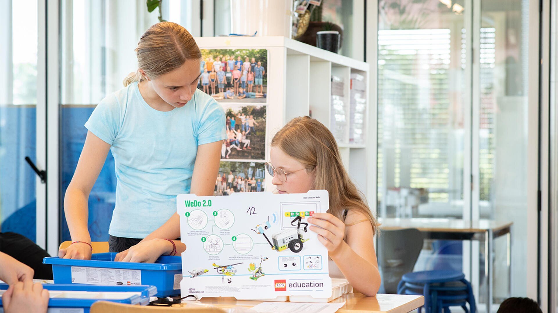 Zwei Mädchen arbeiten zusammen an einem Tisch; eine hält eine LEGO Education WeDo 2.0 Robotik-Anleitung in der Hand, während die andere einen blauen Behälter mit Bauteilen durchsucht. Im Hintergrund sind Regale und die Dekoration des Klassenzimmers zu sehen.
