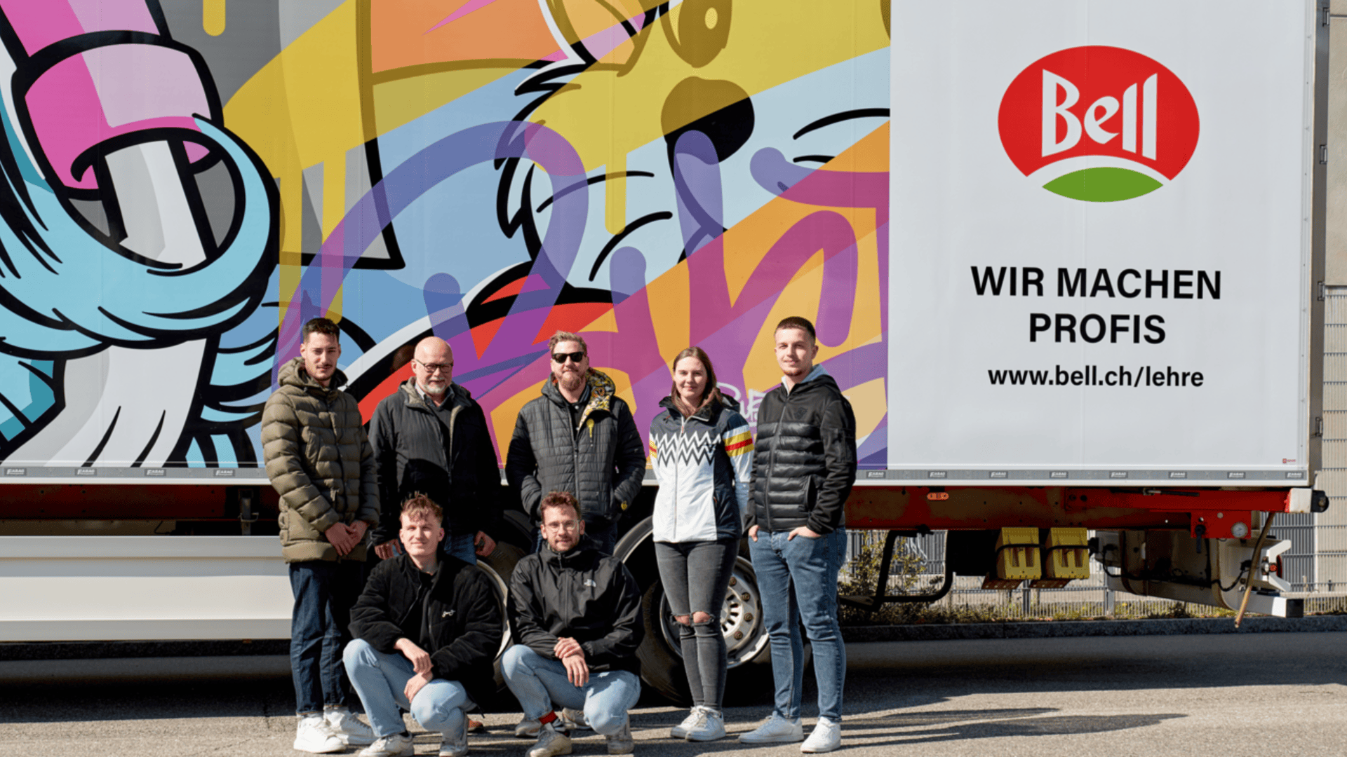Sechs Personen posieren vor einem farbenfrohen Lastwagen mit dem Bell-Logo und dem deutschen Text Wir machen Profis sowie einem Website-Link. Der Lkw ist mit einer großen, lebhaften Tierillustration versehen.