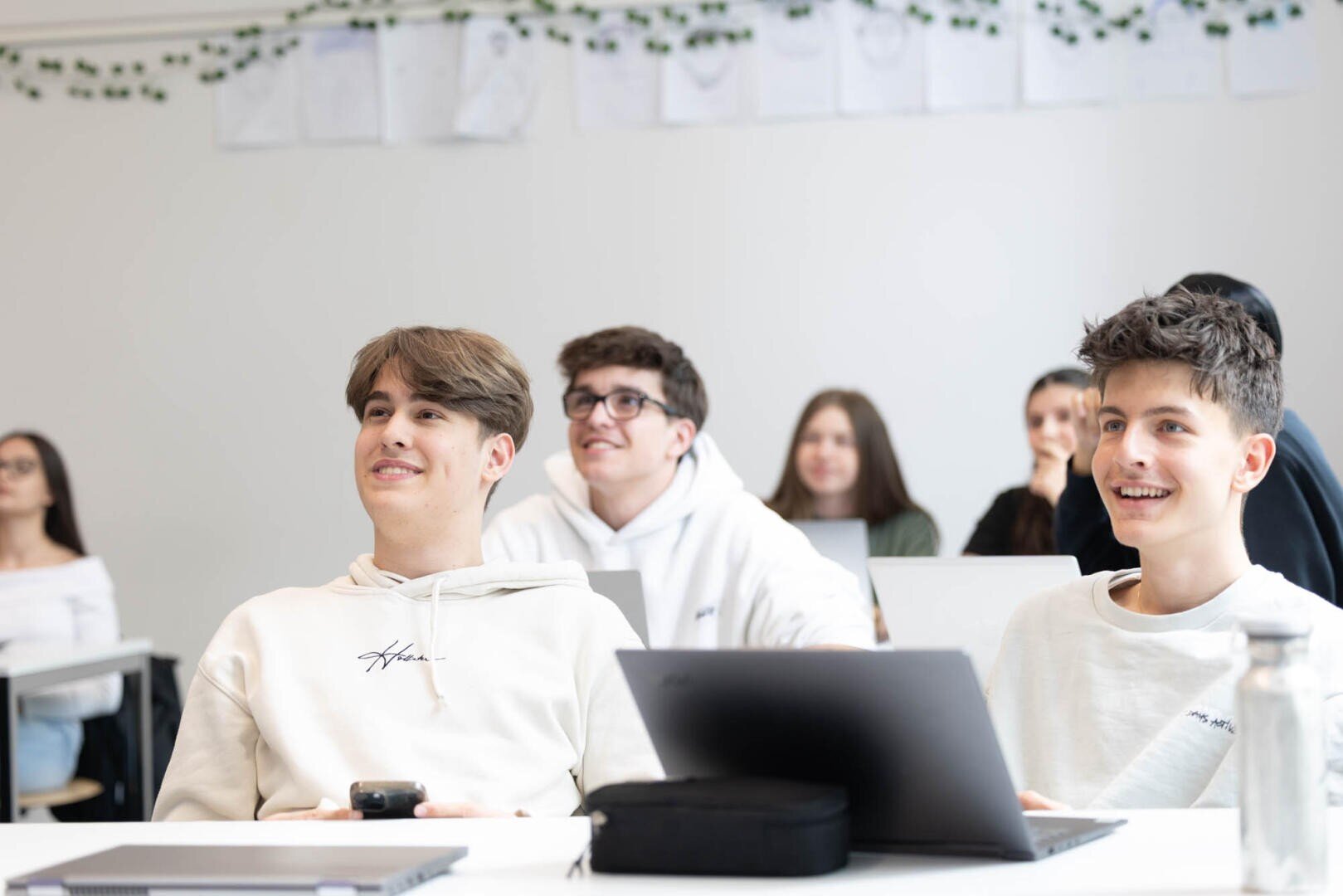 Eine Gruppe von Schülerinnen und Schülern, meist Teenager, sitzt in einem hellen Klassenzimmer an den Tischen, lächelt und blickt nach vorne. Laptops und Notebooks liegen auf ihren Tischen. An die weiße Wand hinter ihnen sind Zeichnungen gepinnt.
