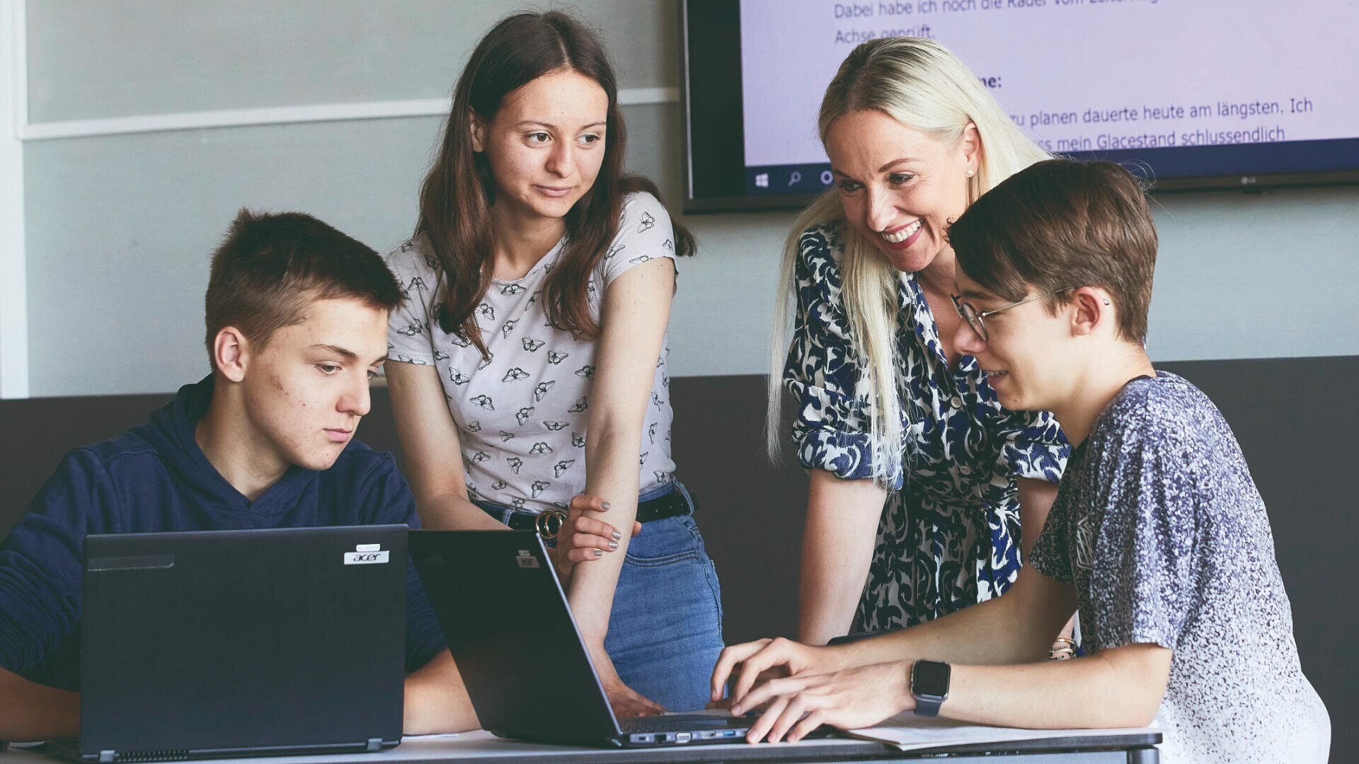Vier Personen, drei junge Erwachsene und ein älterer Erwachsener, sitzen um zwei Laptops herum an einem Tisch, lächeln und diskutieren gemeinsam etwas. Im Hintergrund ist ein großer Bildschirm mit Text zu sehen.
