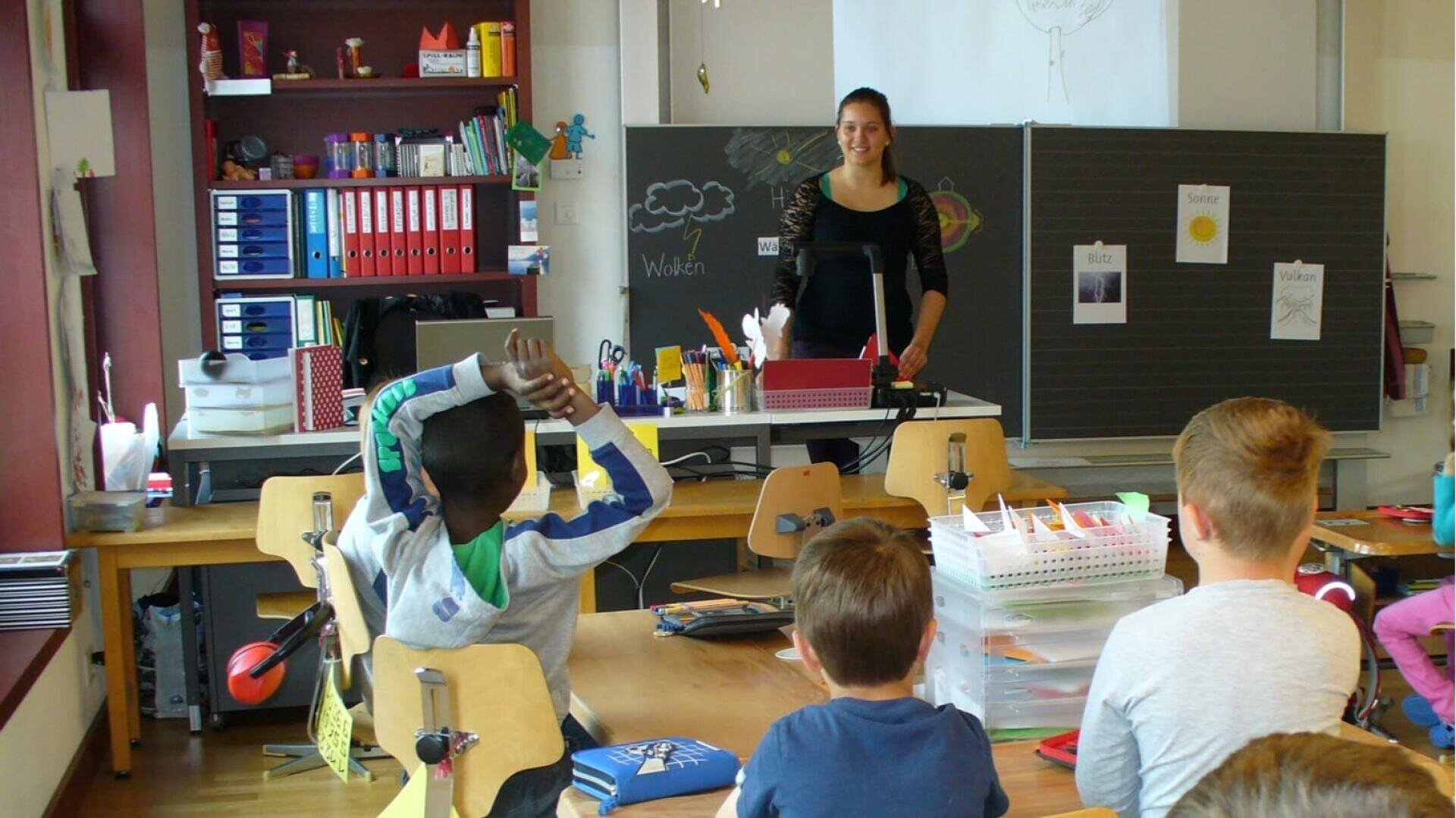 Eine Lehrerin steht vorne in einem Klassenzimmer und lächelt die Schüler an, die an ihren Tischen sitzen. Im Klassenzimmer gibt es Bücherregale, eine Kreidetafel mit Tafeln und verschiedene Schulsachen auf den Tischen.