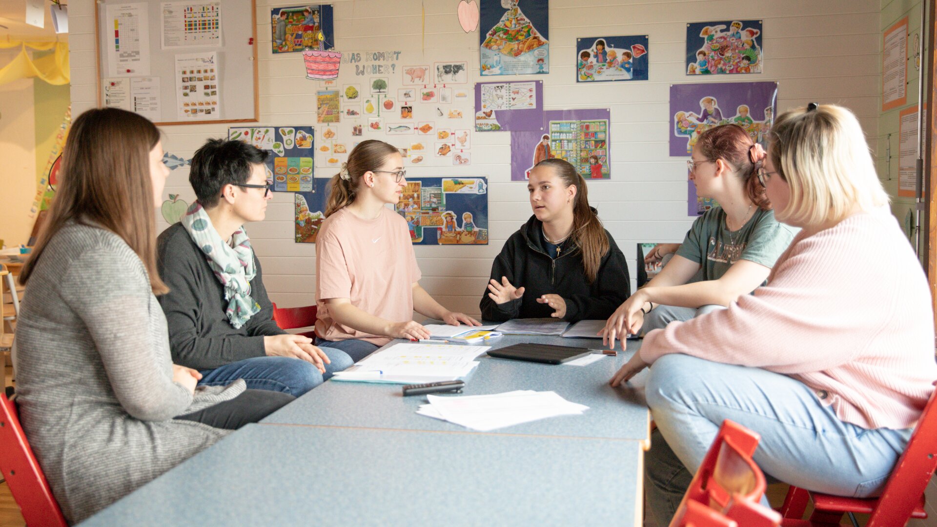 Sechs Frauen sitzen in einem Klassenzimmer um einen Tisch herum und diskutieren, während sie Papiere und Mappen vor sich liegen haben. Bildungsposter und bunte Zeichnungen schmücken die Wand hinter ihnen.
