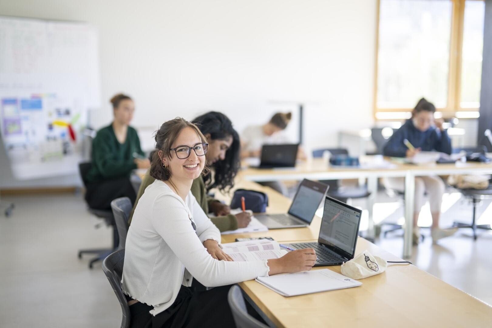 Tourismusstudierende im Klassenzimmer arbeitet am Laptop. Im Hintergrund sitzen mehrere andere Personen an ihren Schreibtischen und sind ganz in ihre Arbeit vertieft.
