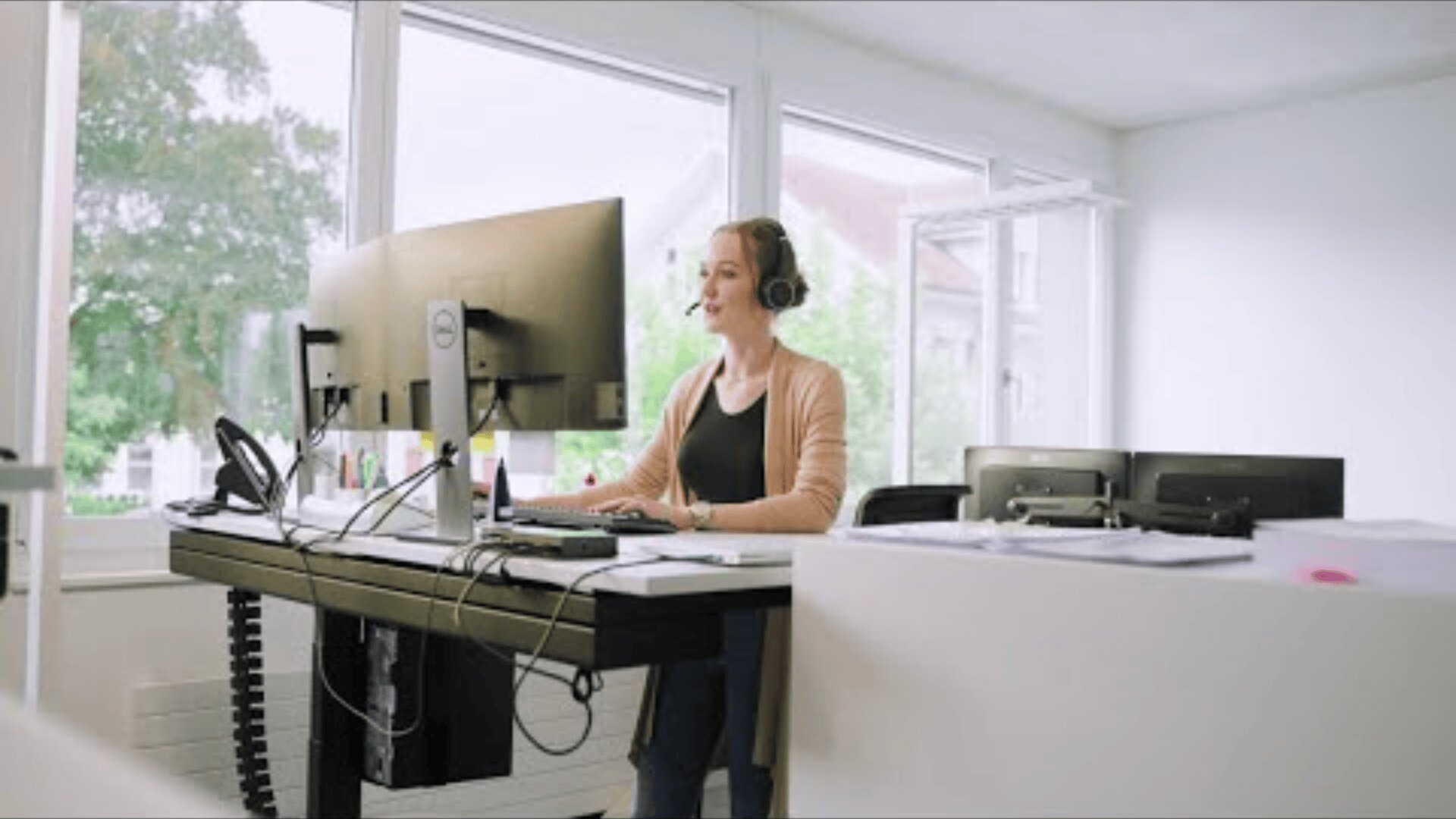 Eine Frau, die ein Headset trägt, sitzt an einem Schreibtisch mit zwei Monitoren in einem hellen, modernen Büro, spricht und arbeitet an ihrem Computer. Große Fenster lassen natürliches Licht herein, und auf dem Schreibtisch liegen Büromaterialien.