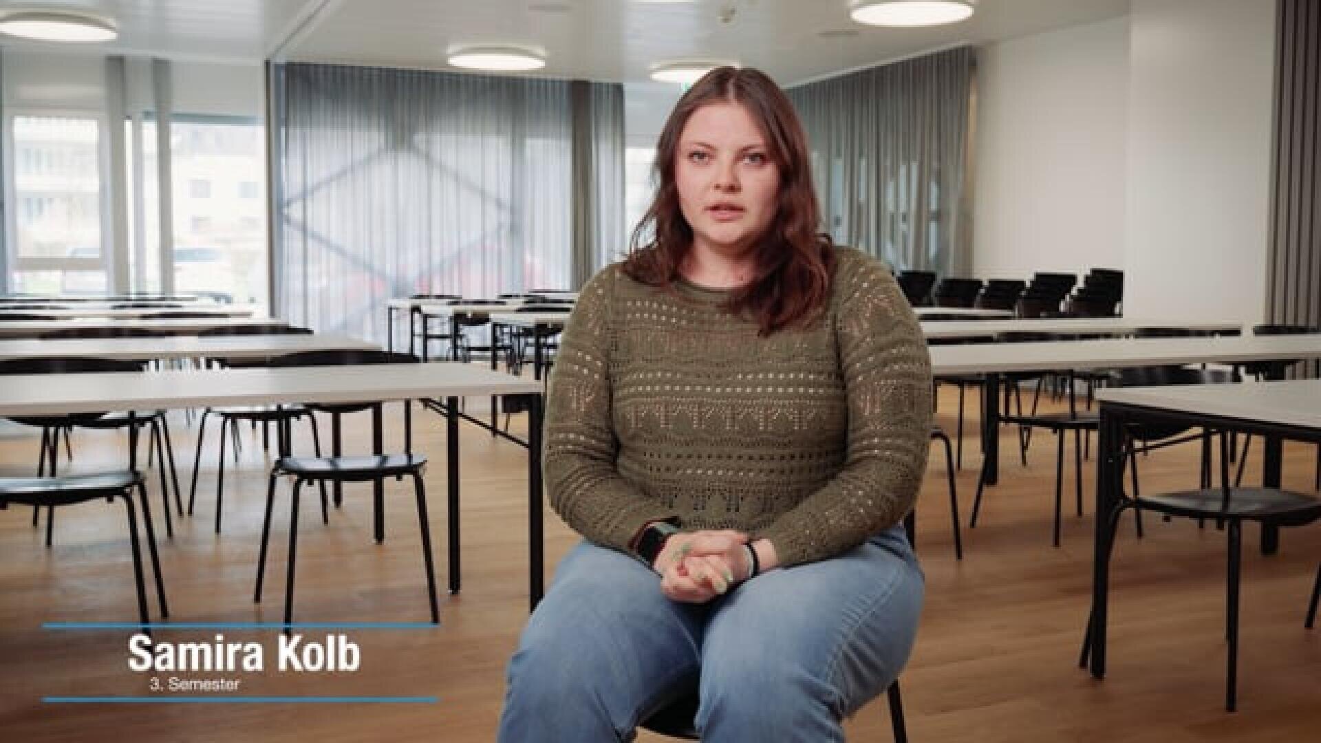Eine Frau mit braunen Haaren sitzt in einem leeren Klassenzimmer; sie trägt einen grünen Pullover und Jeans. Auf dem Bild steht: Samira Kolb, 3. Semester. Im Hintergrund sind große Fenster sowie mehrere Tische und Stühle zu sehen.